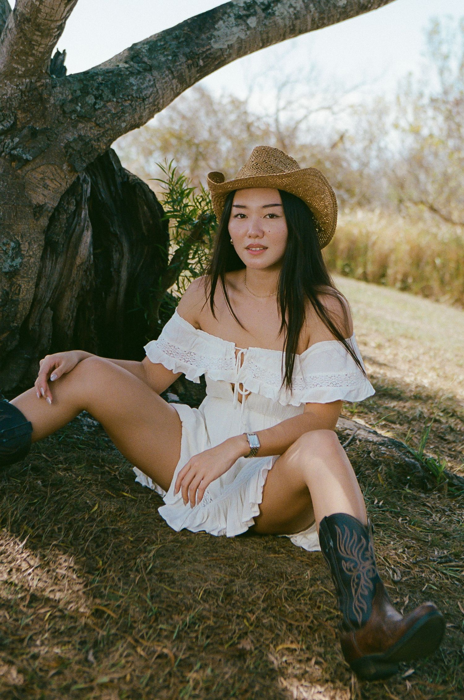 Young woman in white off-shoulder dress and cowboy hat sitting under tree in natural setting.