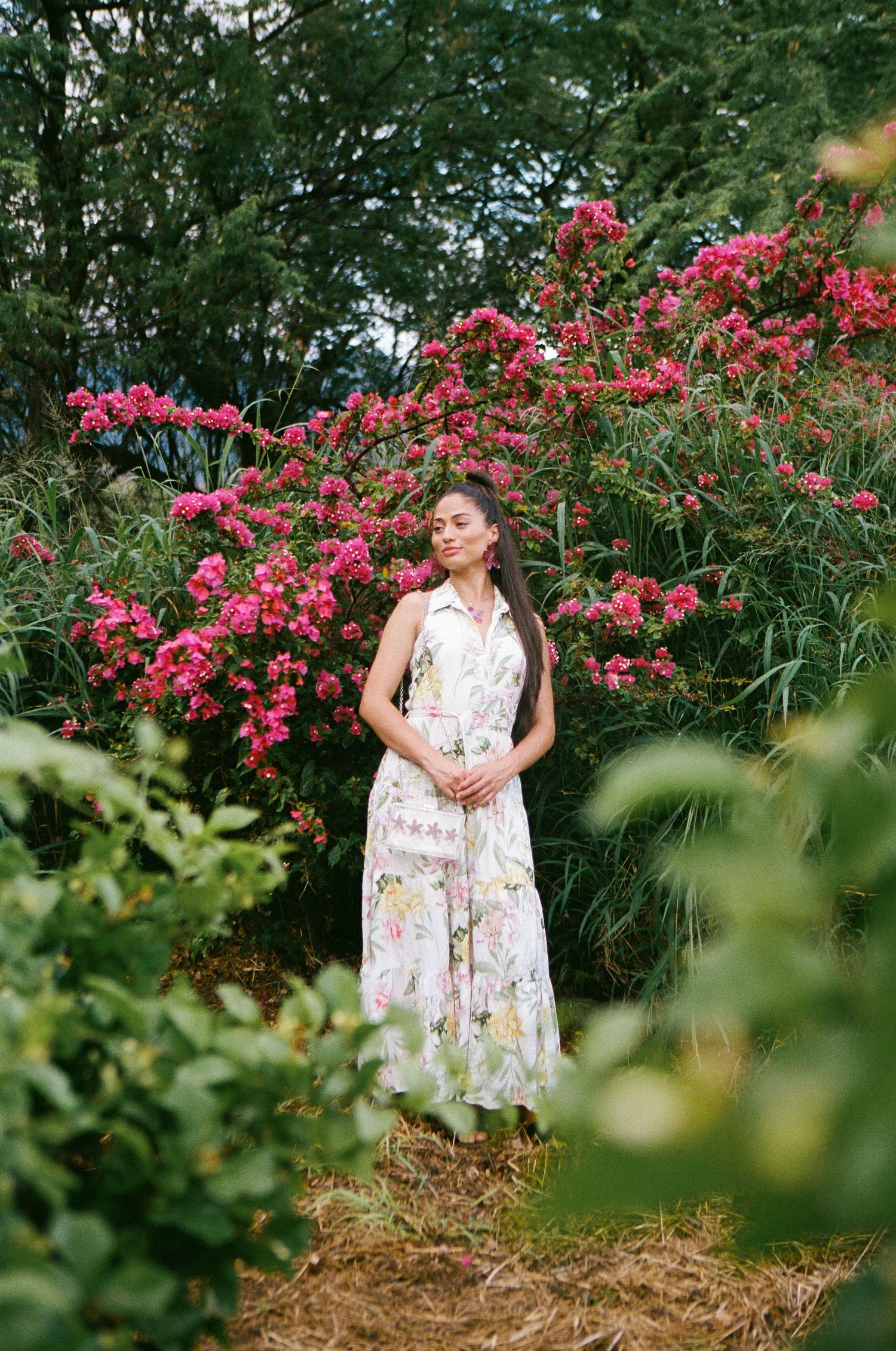 Woman in floral maxi dress standing among pink blooming flowers and lush green garden foliage.