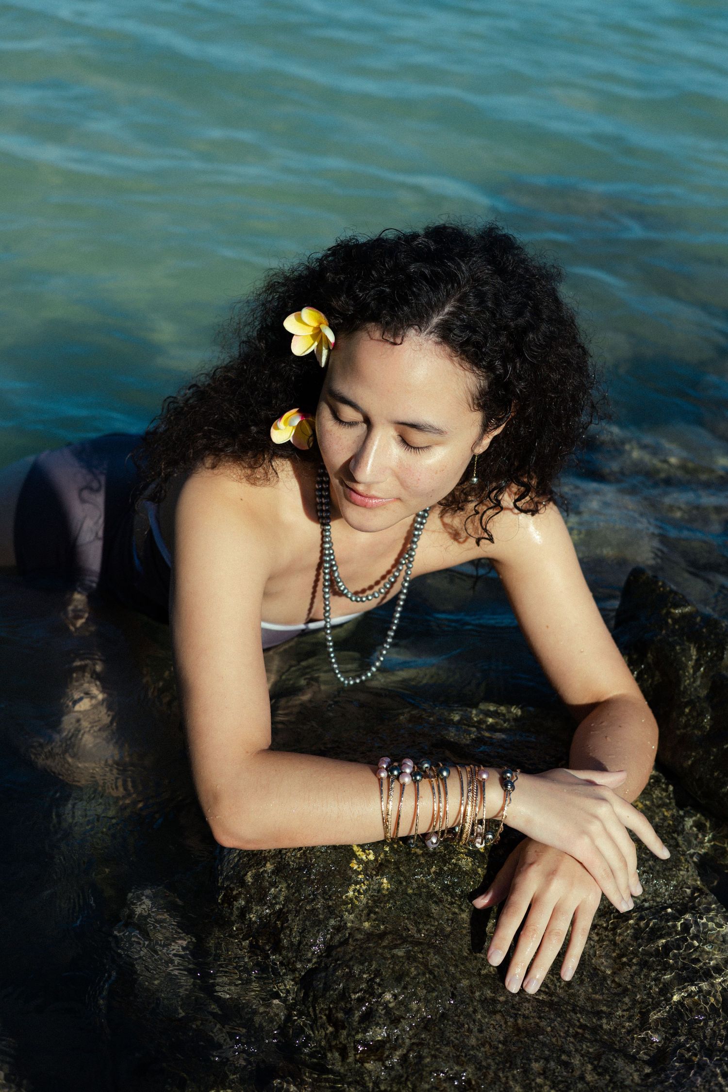Curly-haired woman with flower in hair and beaded necklaces resting on rocks in ocean.