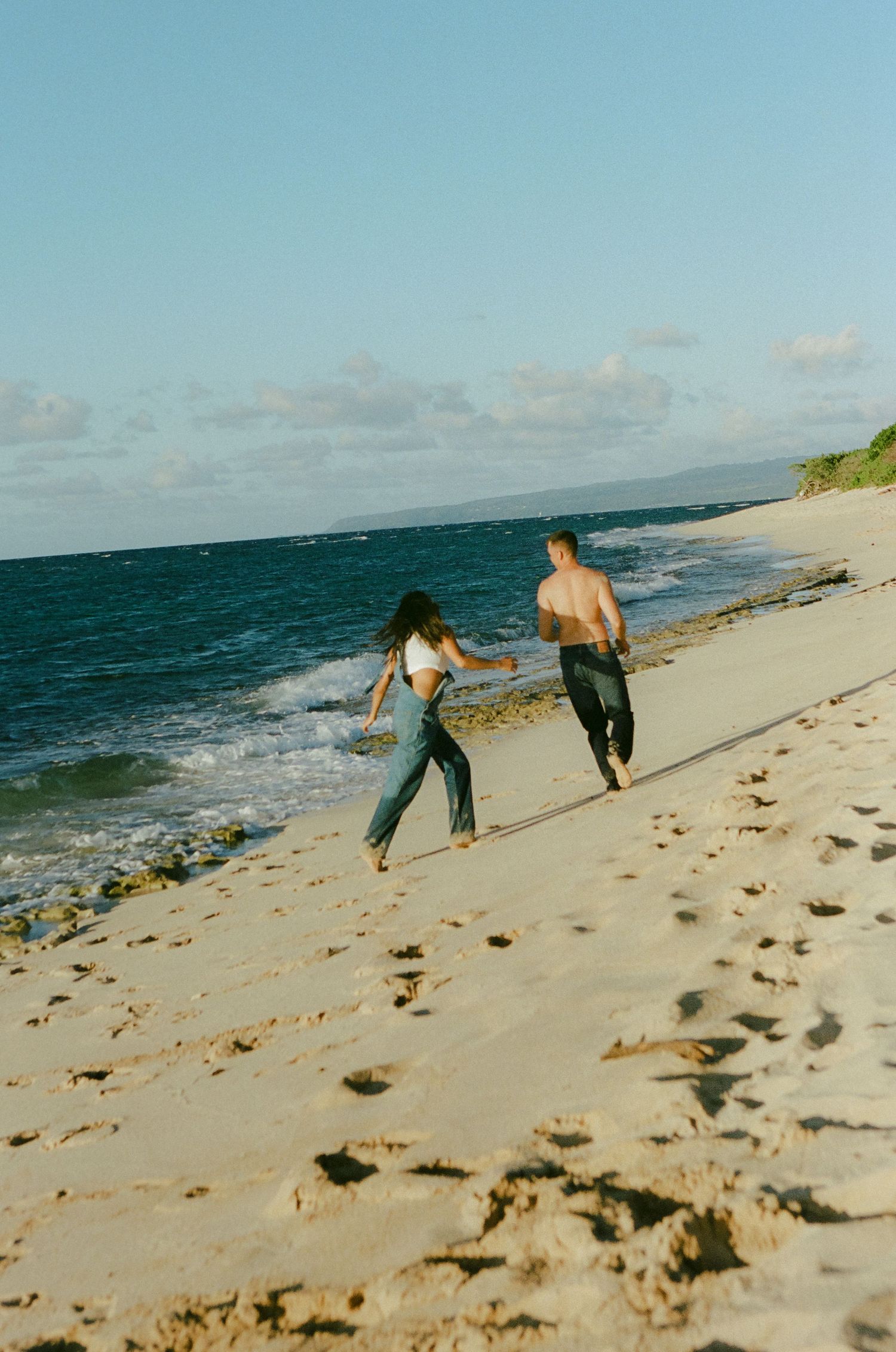 Couple running barefoot along sunny beach shoreline, woman in jeans and crop top, man shirtless.