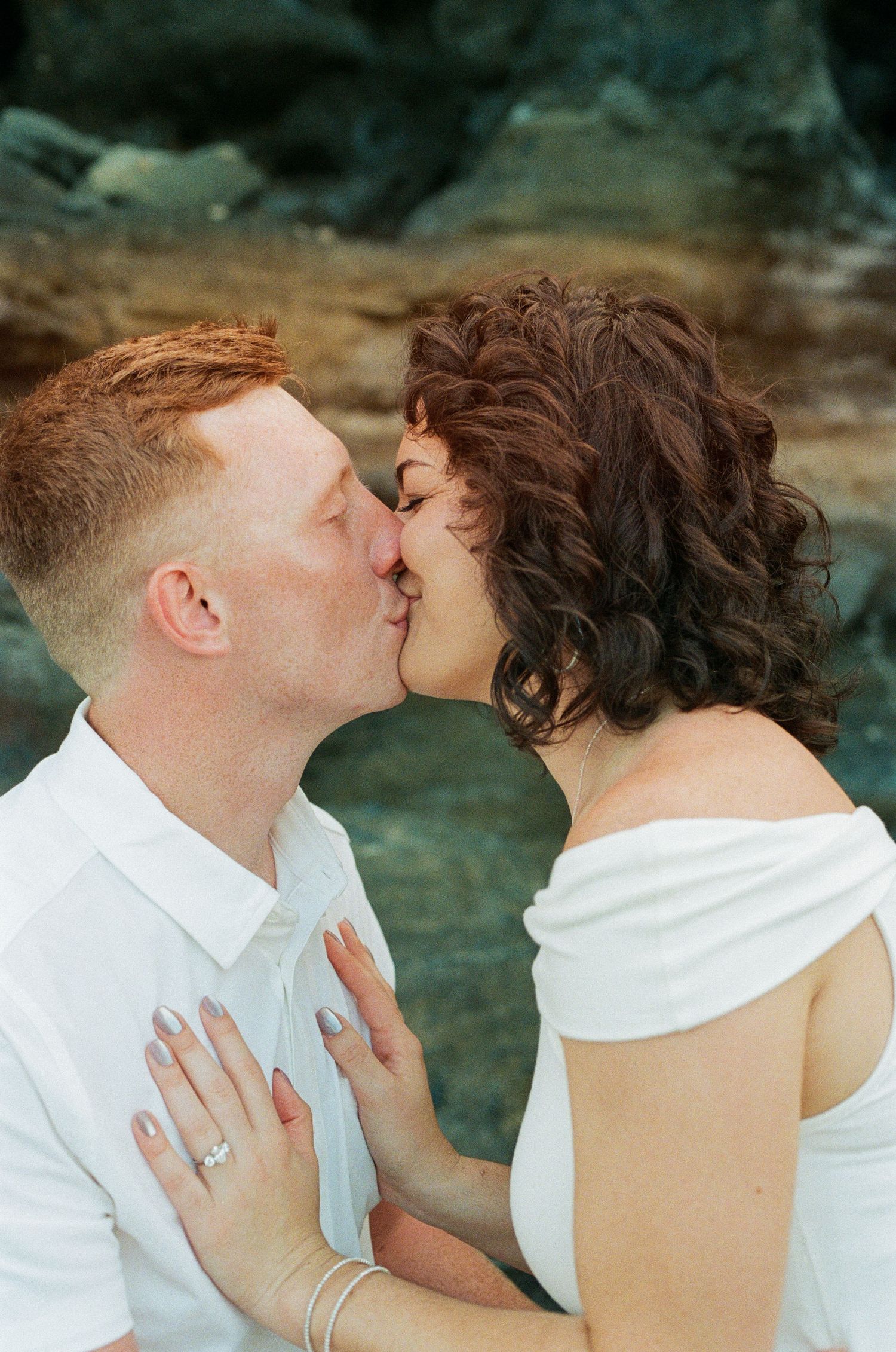 Engaged couple kissing near rocky backdrop, woman in off-shoulder white dress showing diamond ring.