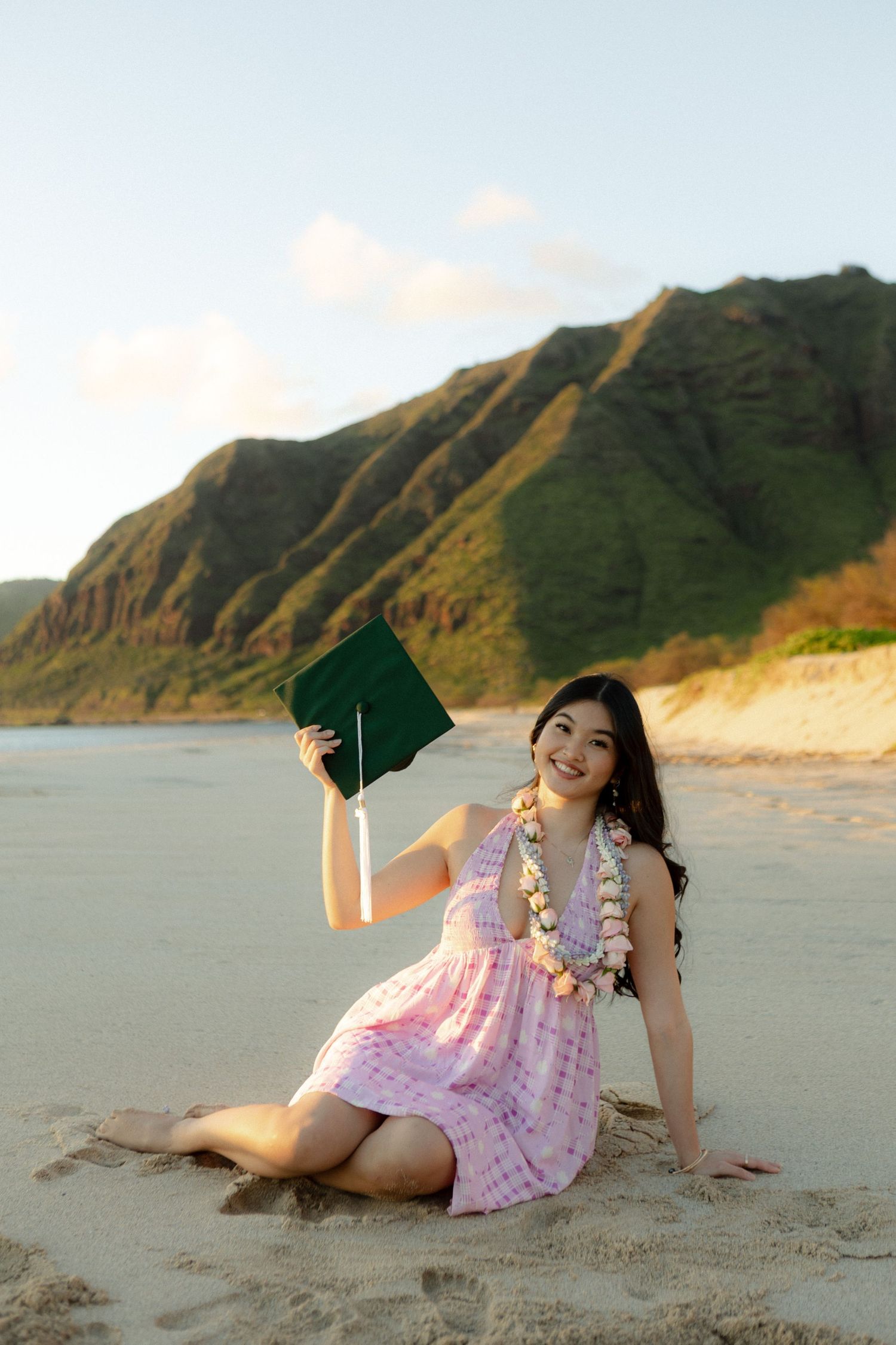 Smiling woman in pink dress holds green graduation cap seated on Hawaiian beach.