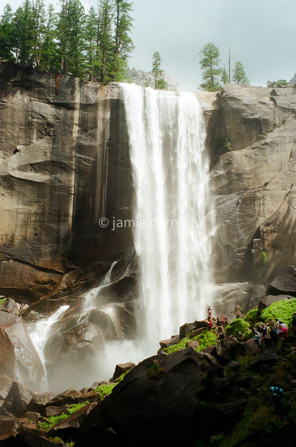 Majestic waterfall cascading down granite cliffs in Yosemite, with hikers on mossy rocks below and pine trees above.