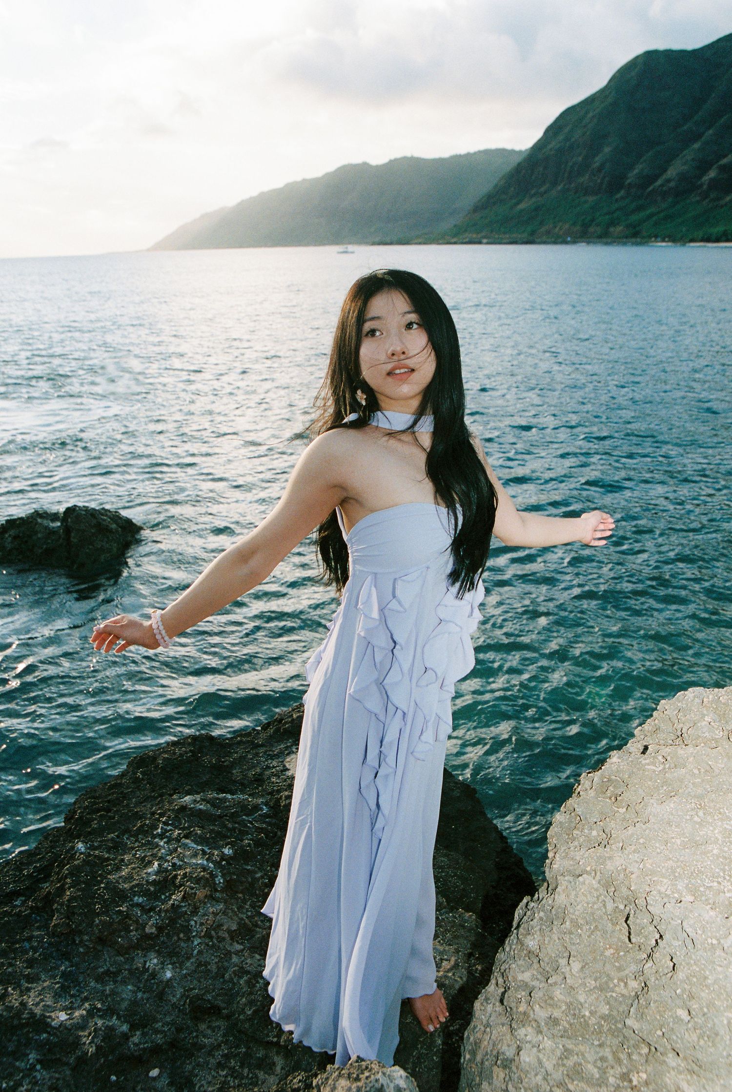 Young woman in a light blue ruffled dress posing on rocky coastline with turquoise ocean and mountains behind her.