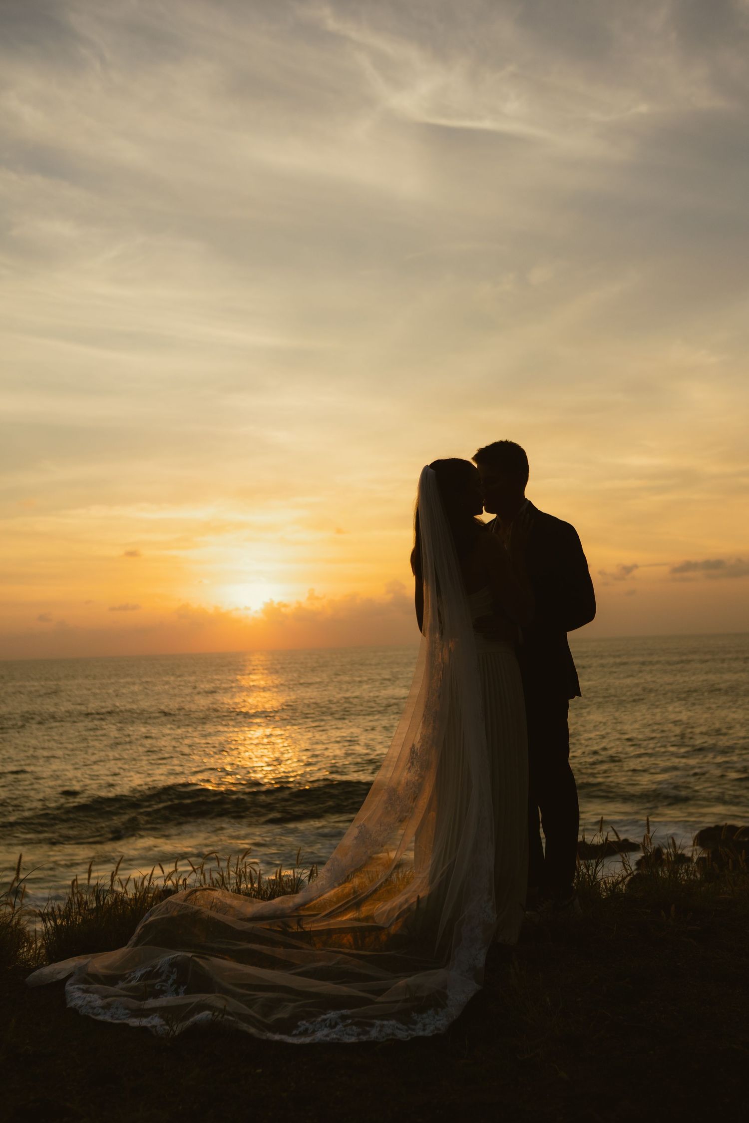 Bride and groom silhouette embracing at sunset over ocean, wedding veil flowing in breeze.
