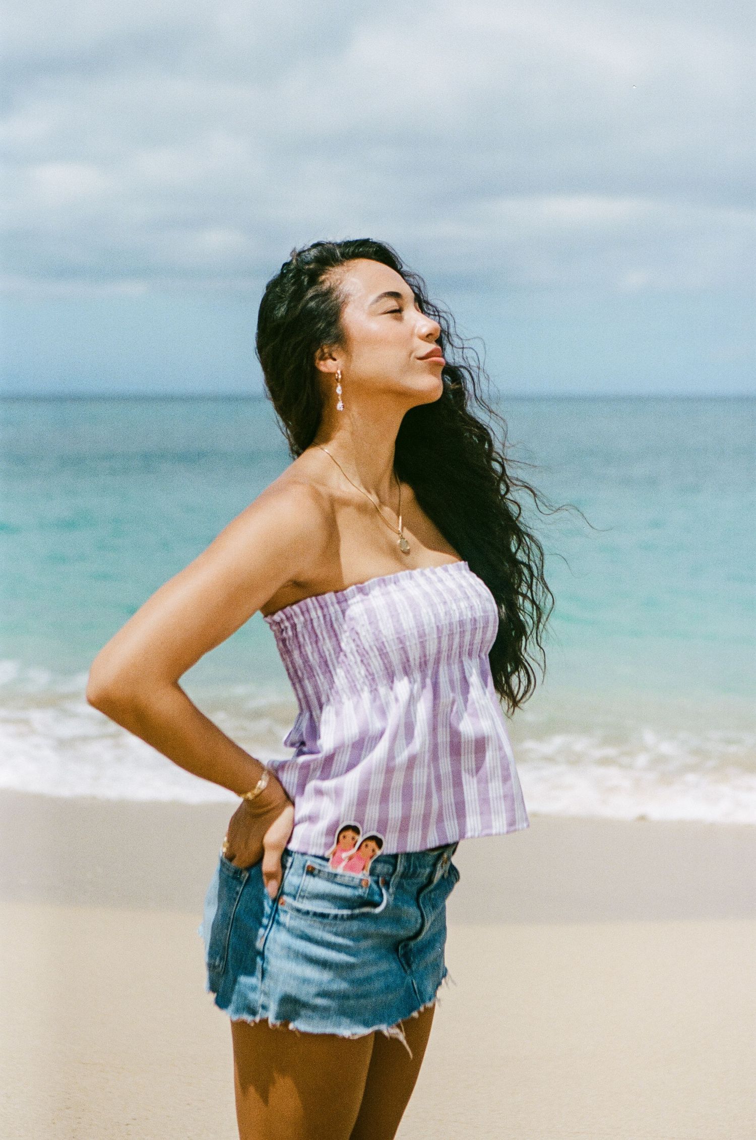 Woman in purple gingham strapless top and denim skirt posing confidently on a tropical beach shoreline.