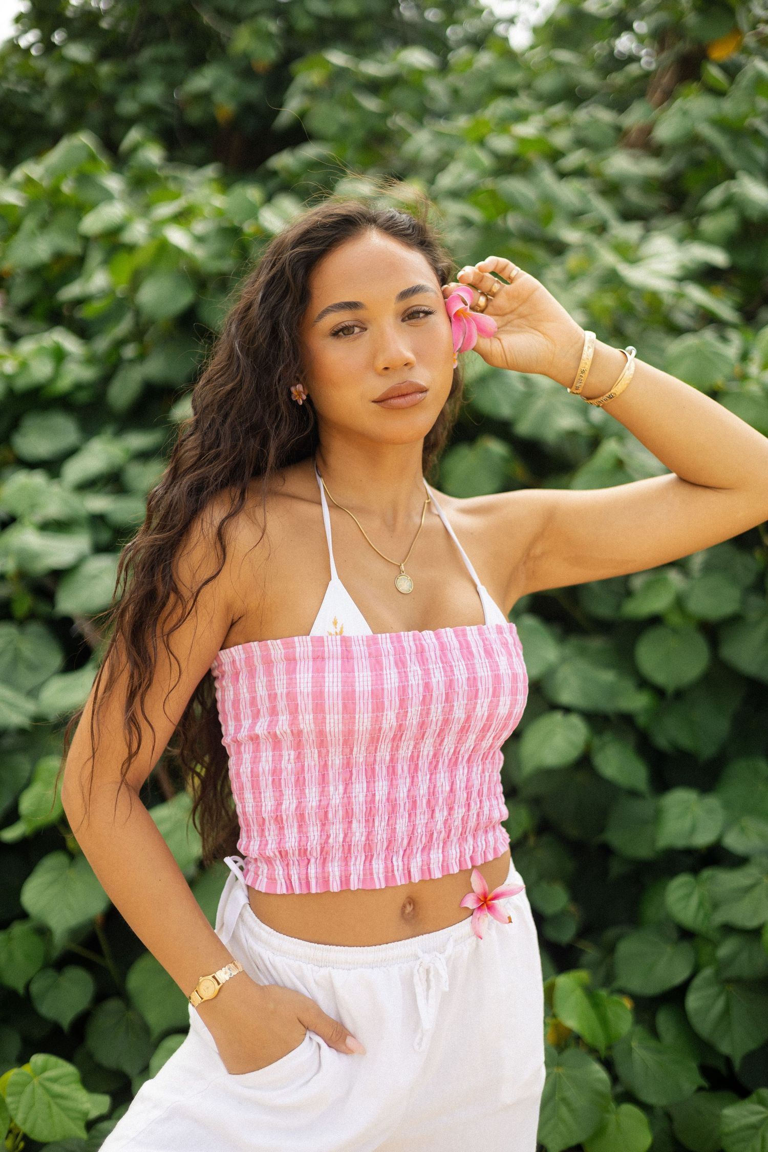 Woman in pink striped halter crop top and white pants posing in front of vibrant green tropical leaves.