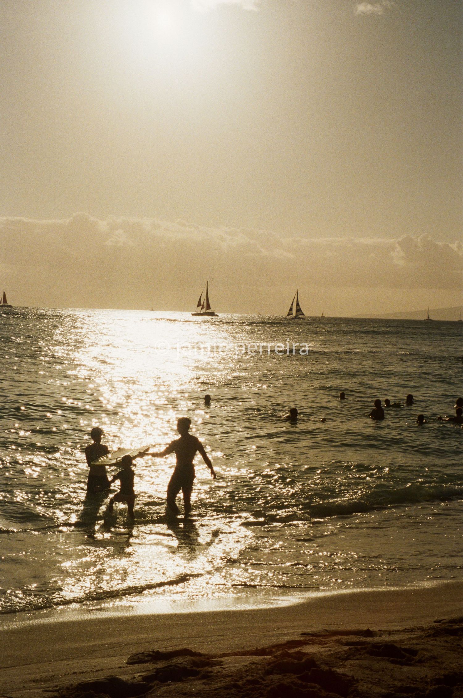 Silhouettes of people playing in ocean waves at sunset with sailboats on the horizon during golden hour.