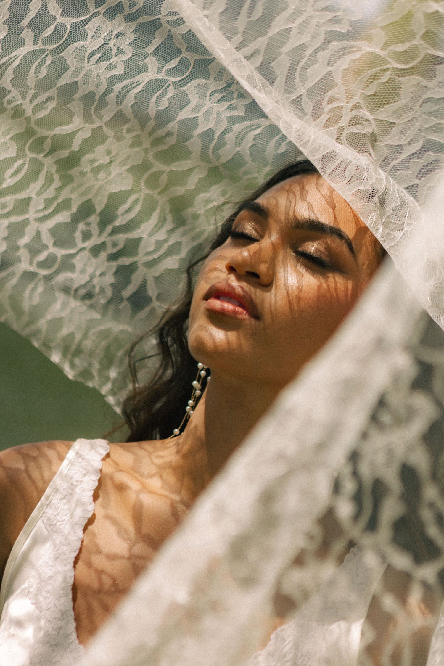 Close-up portrait of bride with closed eyes beneath lace veil with sunlight and shadow patterns.