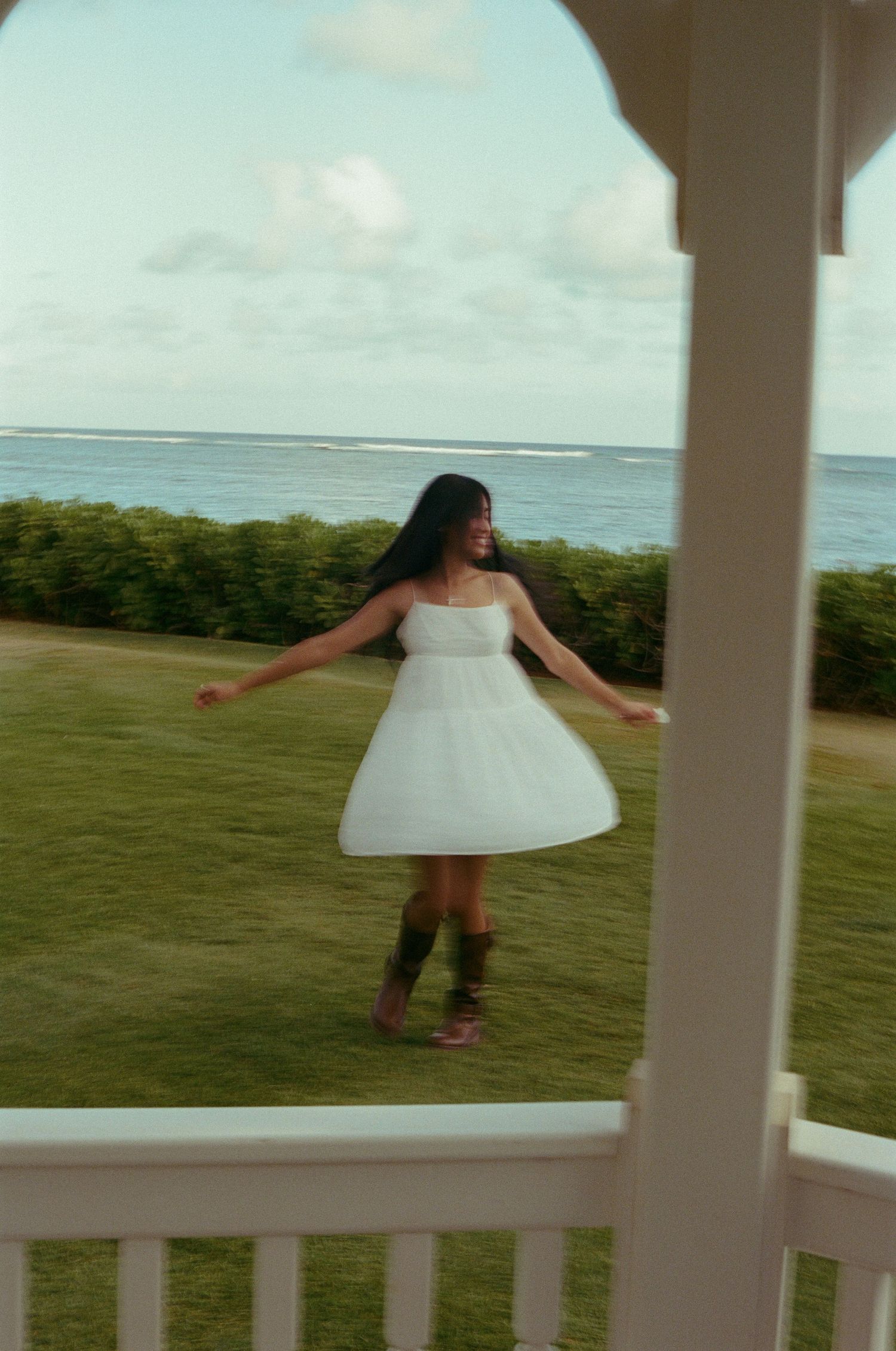 Woman in white mini dress spinning on green lawn with ocean horizon visible in background.
