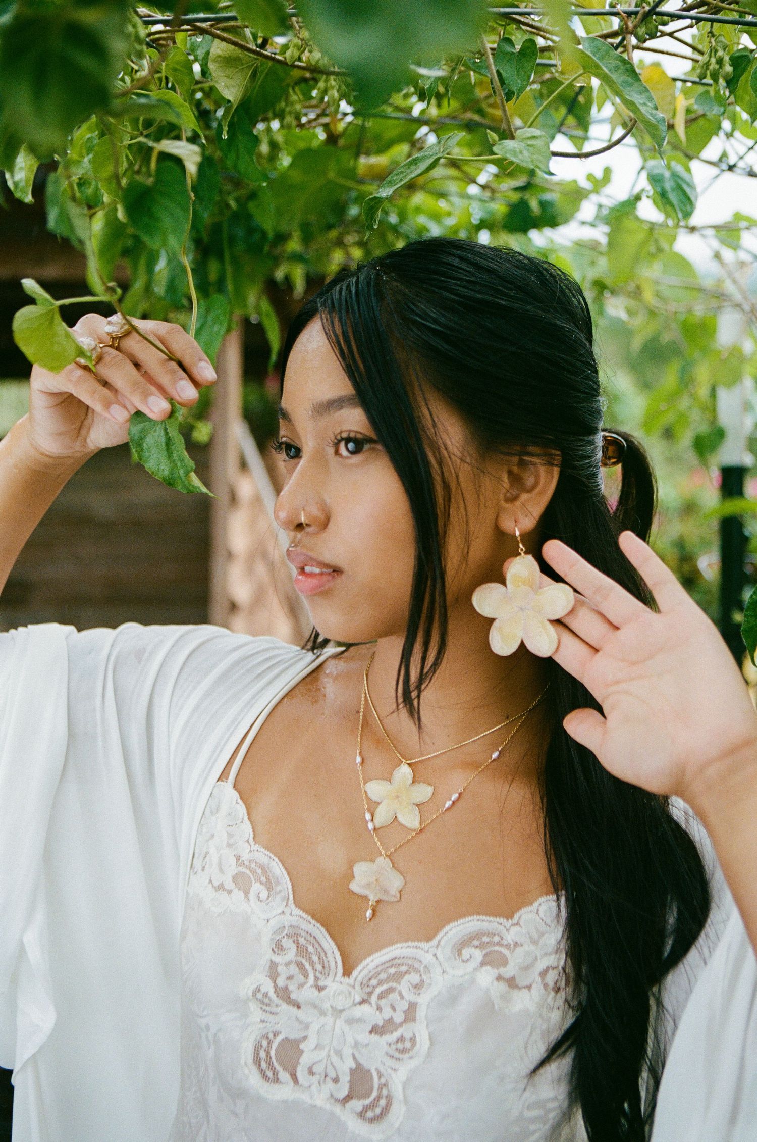 Young woman in white lace dress wearing large flower earrings and ring among green leaves.