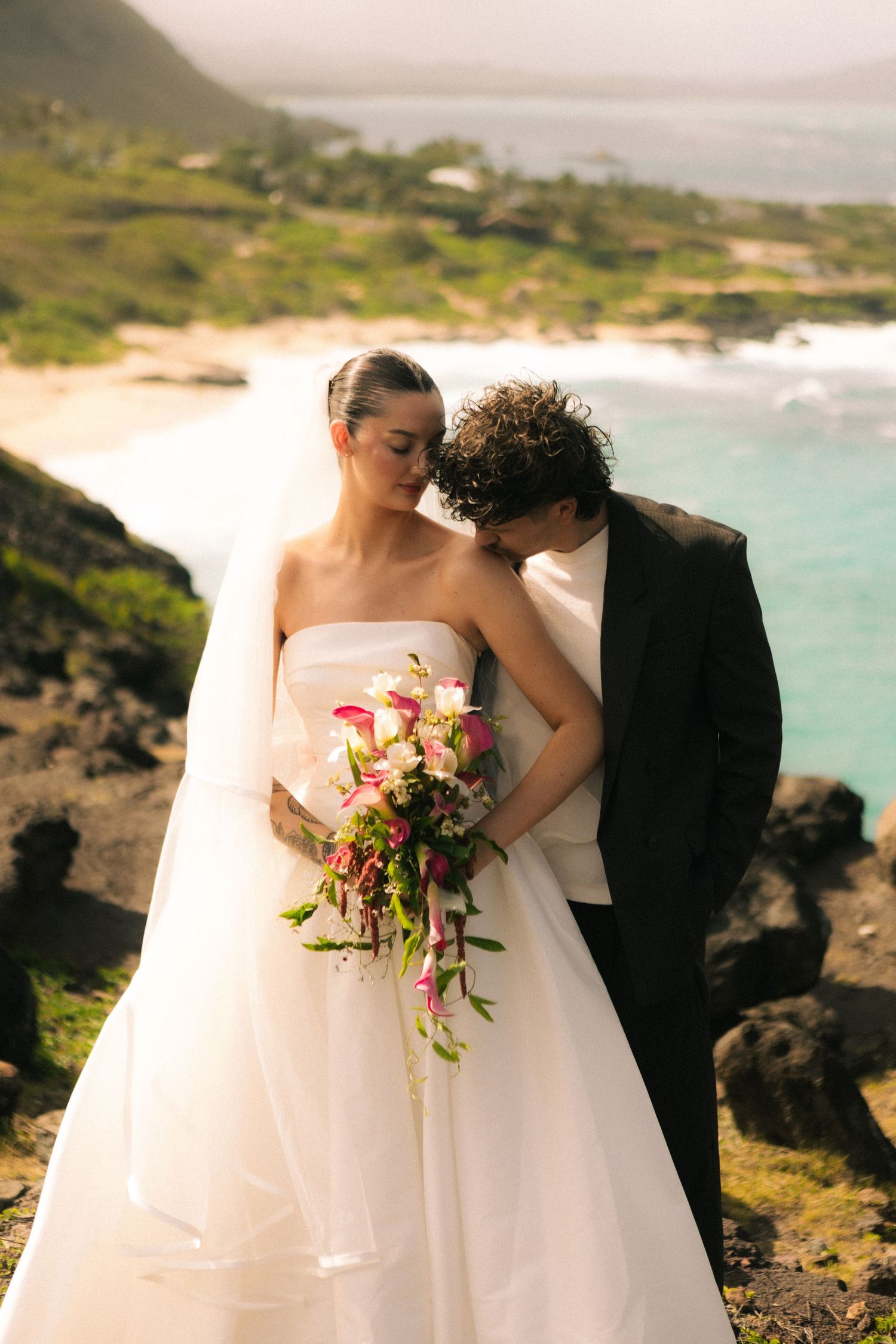 Bride in white gown holding pink bouquet with groom in black tuxedo posing on rocky Hawaiian coastal cliffside.