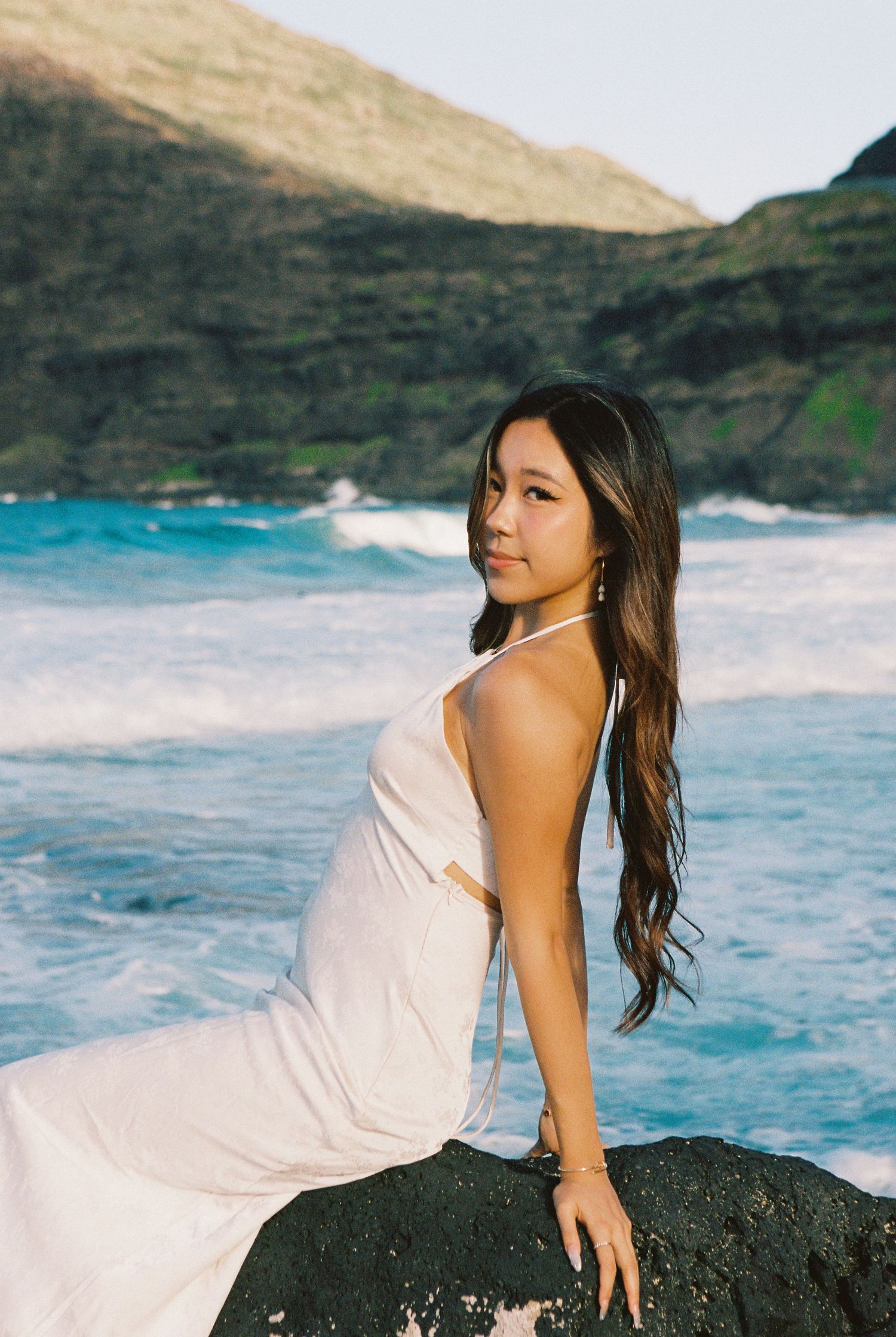 A young woman in a white halter dress sits on a rock by a turquoise ocean with green cliffs behind her.