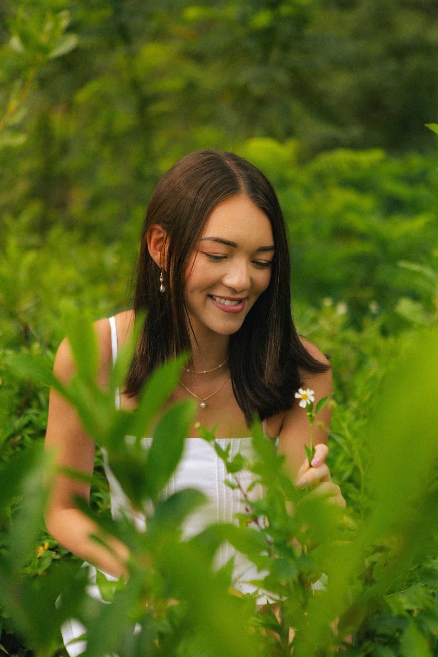 Smiling young woman in white top surrounded by lush green foliage holding a flower.