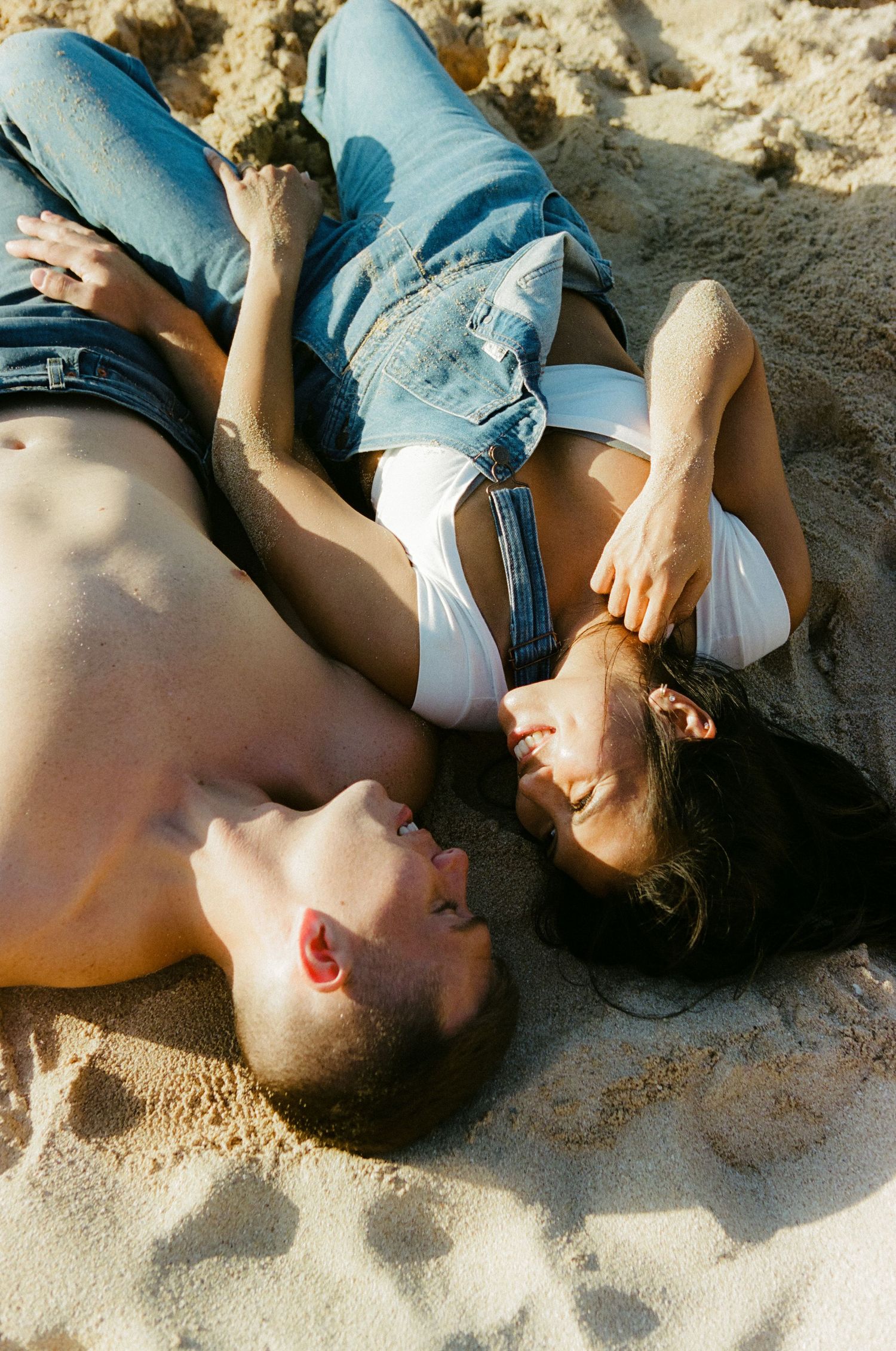 Couple lying in sand laughing together, wearing jeans, enjoying a sunny beach day.