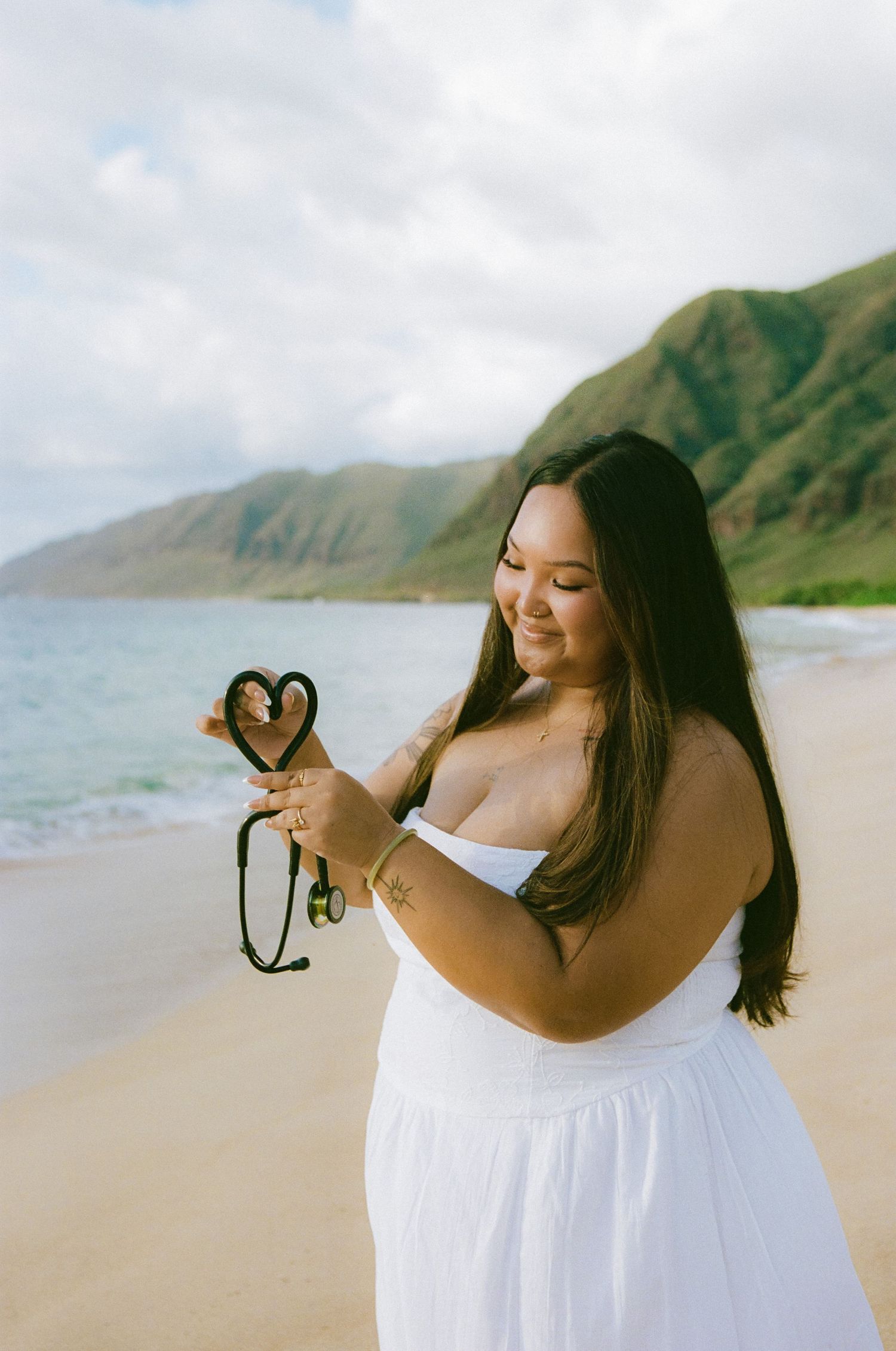 Smiling woman in white dress on beach holding camera strap in heart shape with mountains behind.