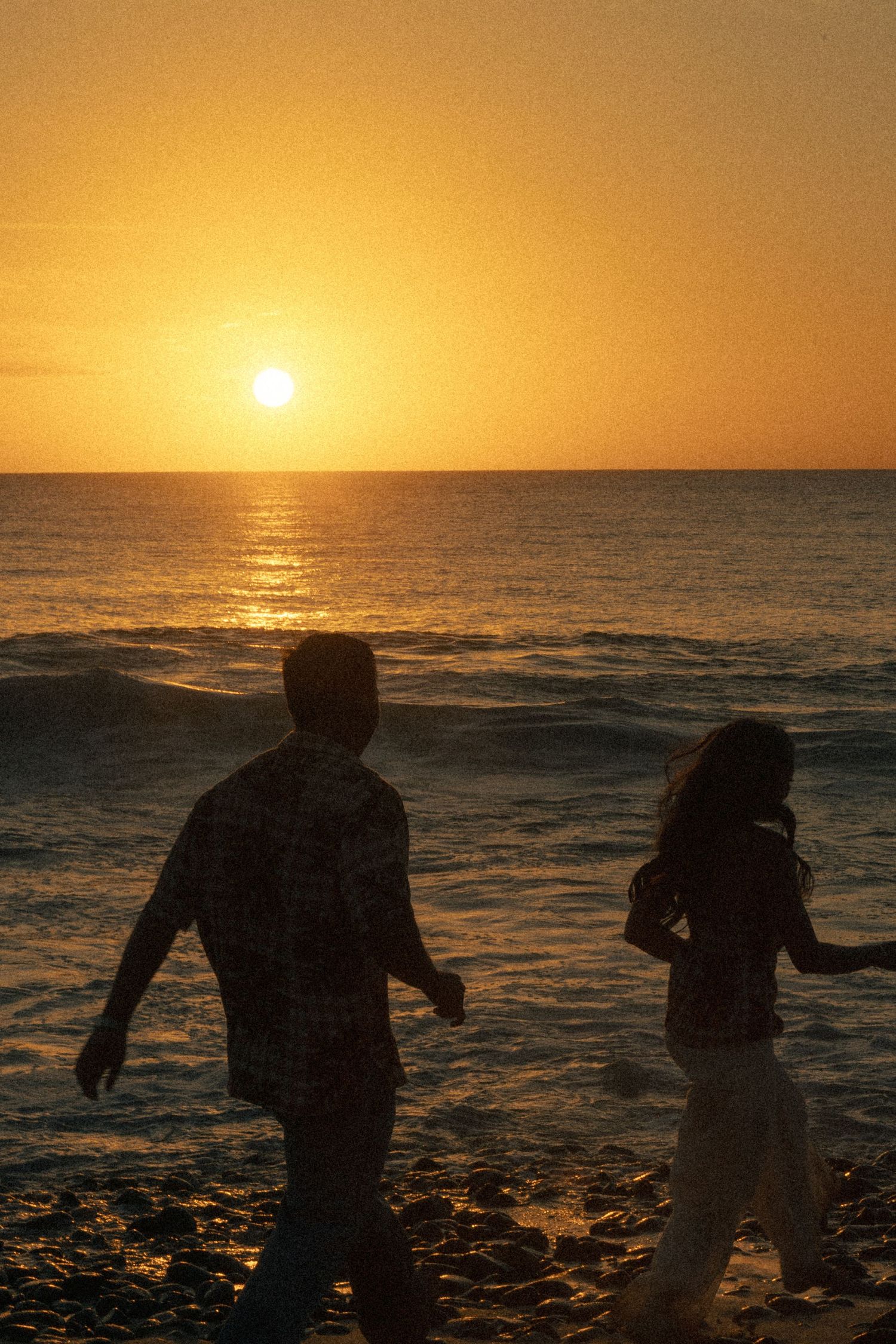 Two silhouetted figures walk along a beach at golden sunset with waves and glowing orange sky over the ocean.