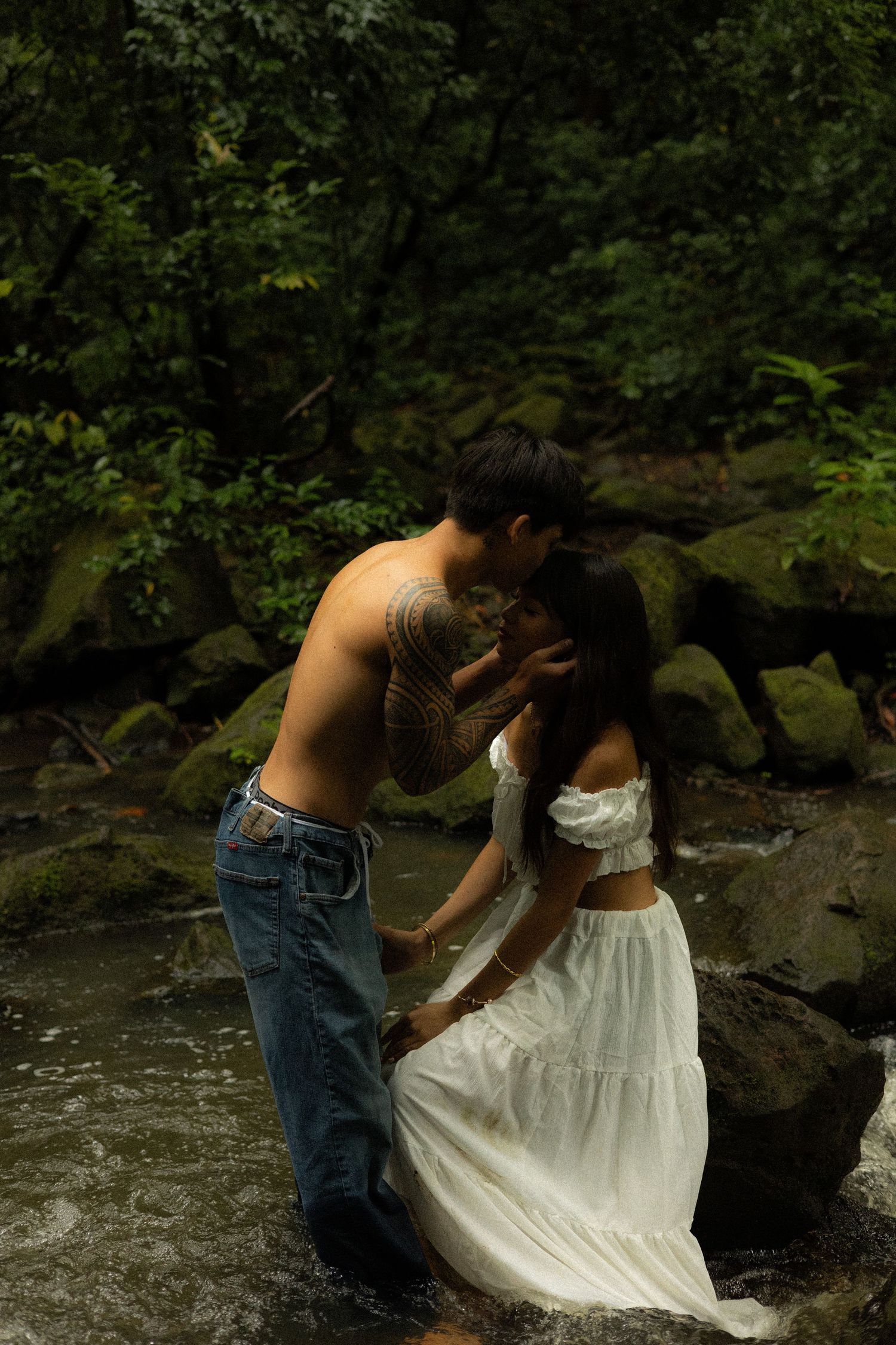 Shirtless tattooed man kissing woman in white dress while standing in a lush tropical forest stream.