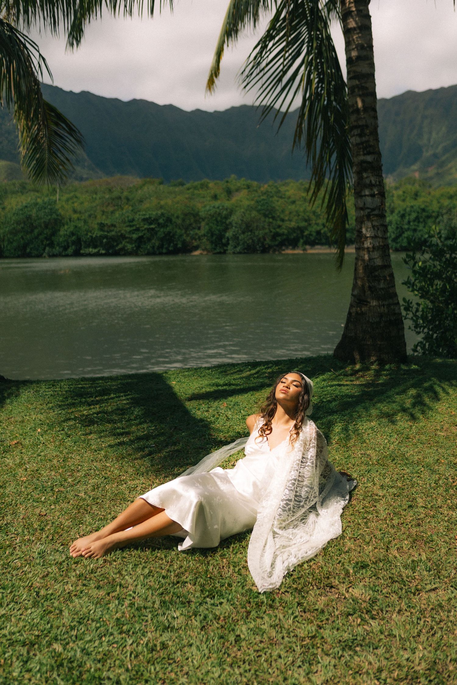 Bride in white dress relaxing under palm tree by tranquil lake with mountain views in Hawaii.
