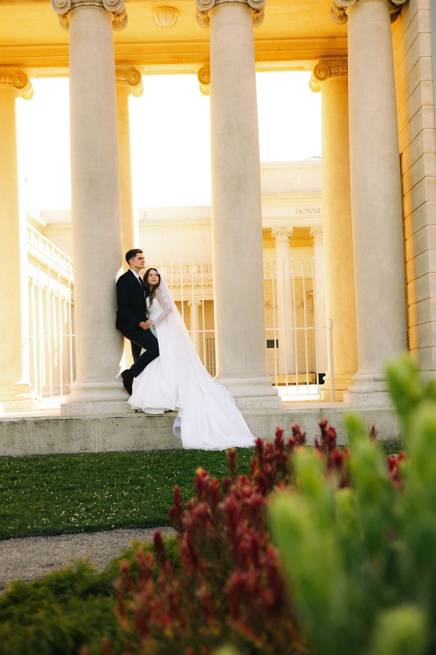Bride and groom posing on steps of elegant columned building in wedding attire, long white veil trailing gracefully.