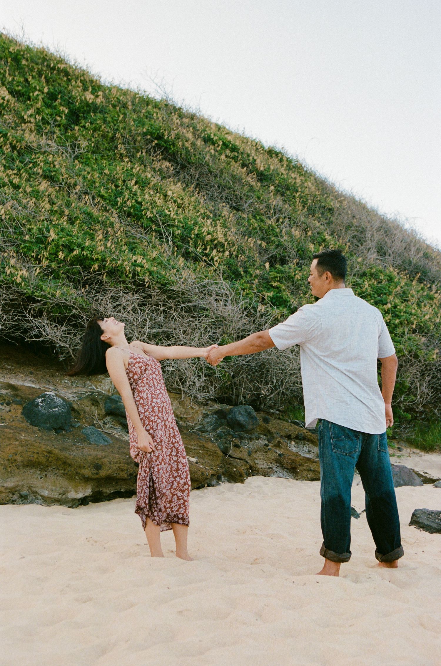 A couple holds hands on a sandy beach, woman leaning back gracefully near a green hillside.