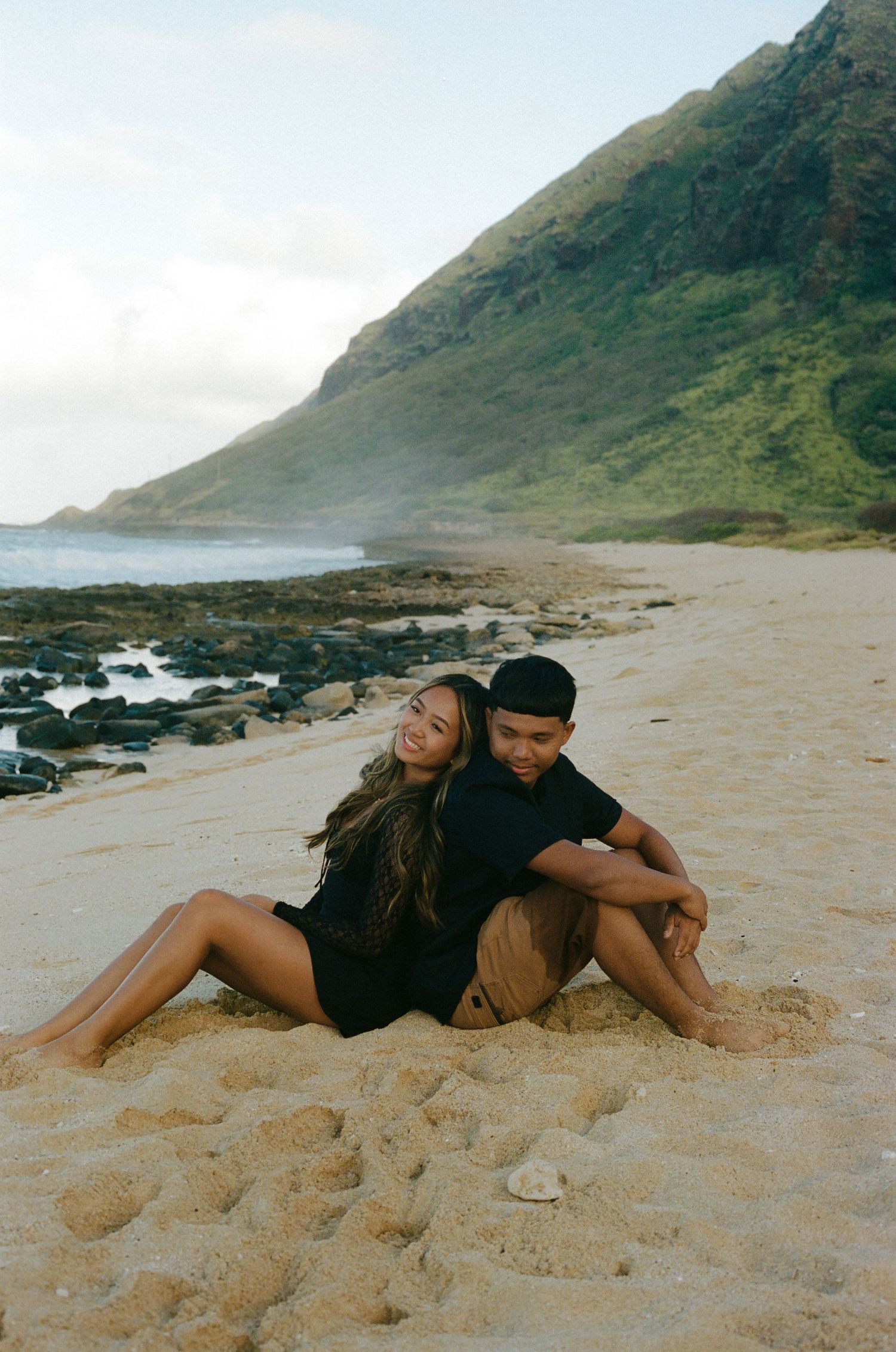 Couple sitting back to back on a sandy beach with mountain backdrop.