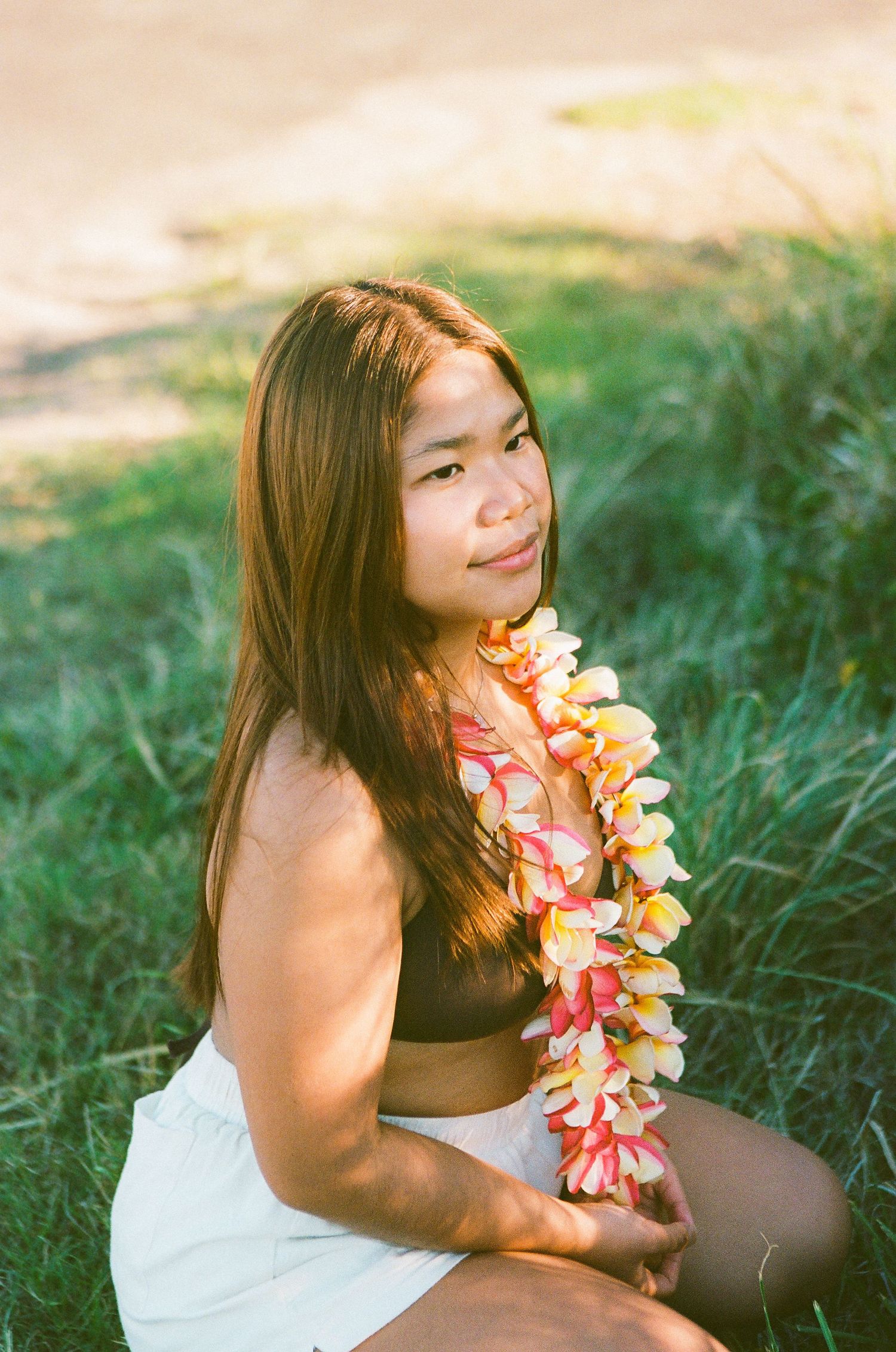 Young woman sitting on grass wearing a floral lei necklace with long brown hair, smiling outdoors.
