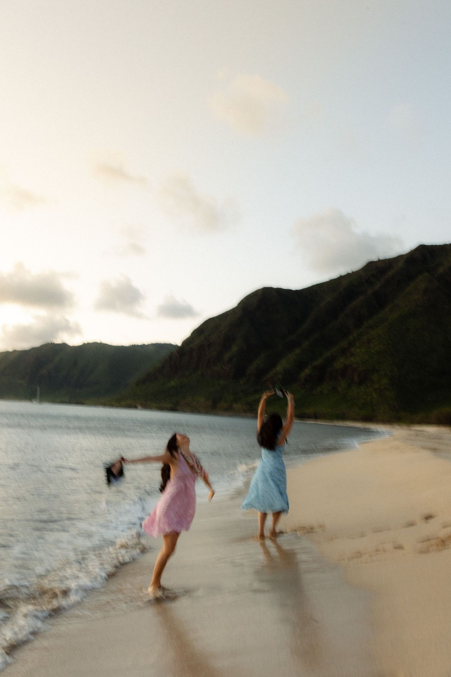 Two women in colorful dresses dance joyfully on a Hawaiian beach at sunset.