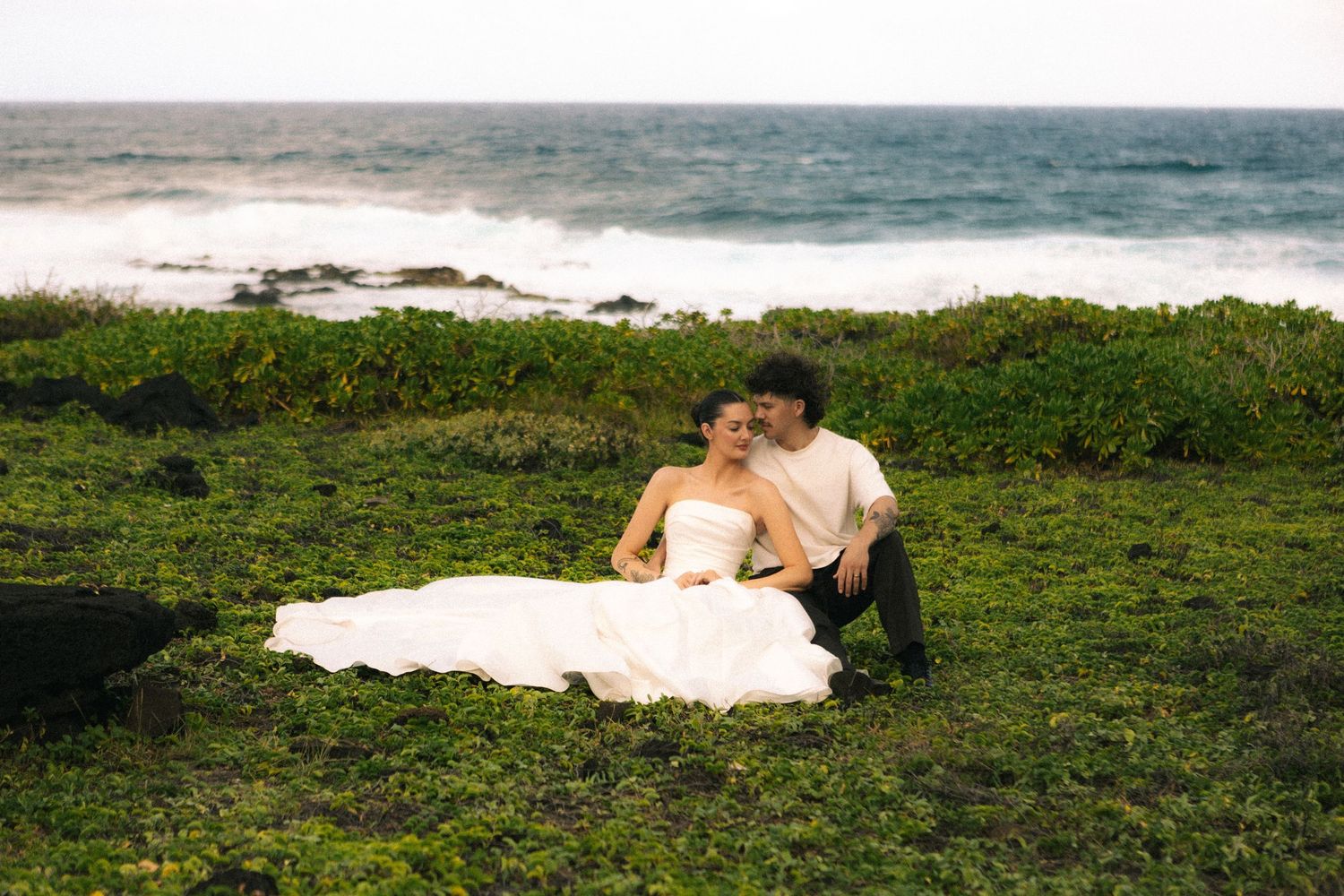 Couple in wedding attire sitting on green coastal cliffs with ocean waves in the background.