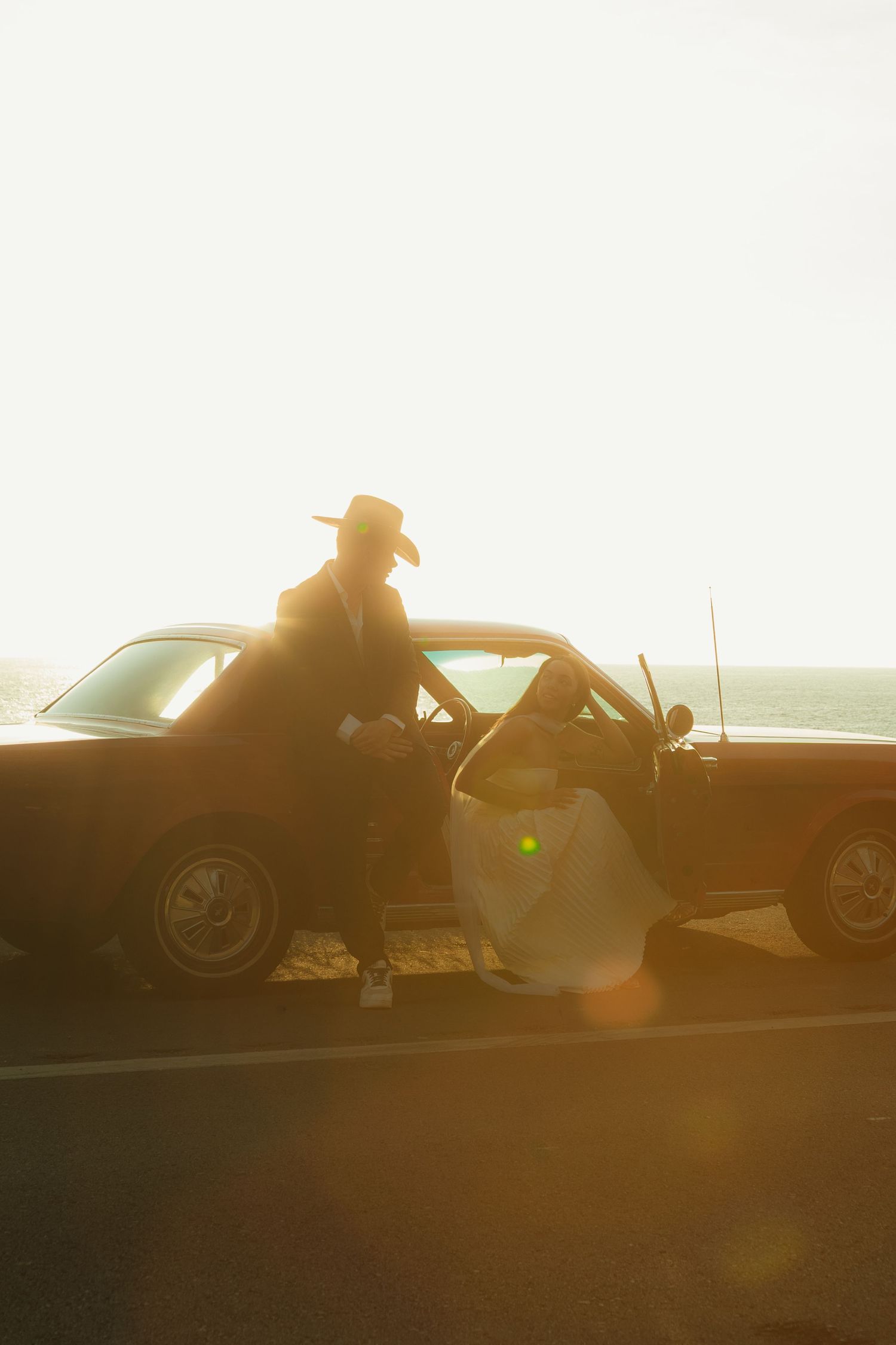Couple in vintage car at golden sunset by the ocean, man in hat leaning on classic car door in warm sunlight.