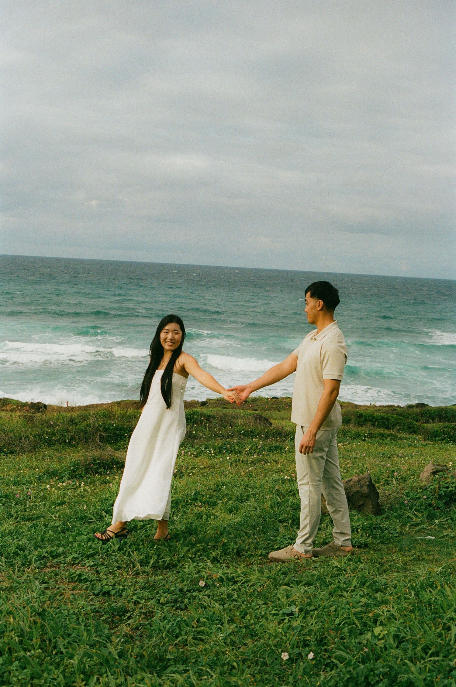 Couple holding hands facing each other on grassy coastal cliff with turquoise ocean waves behind.
