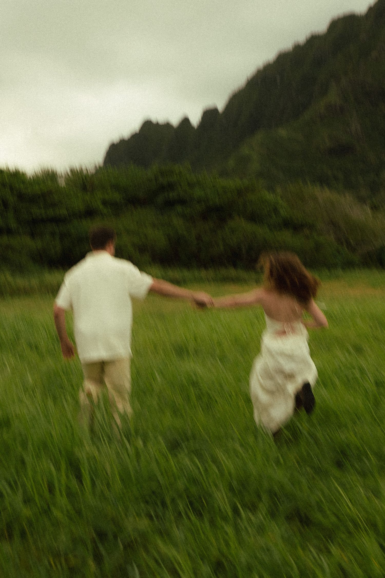 Couple holding hands running through green meadow with dramatic misty mountain backdrop, motion blur effect.