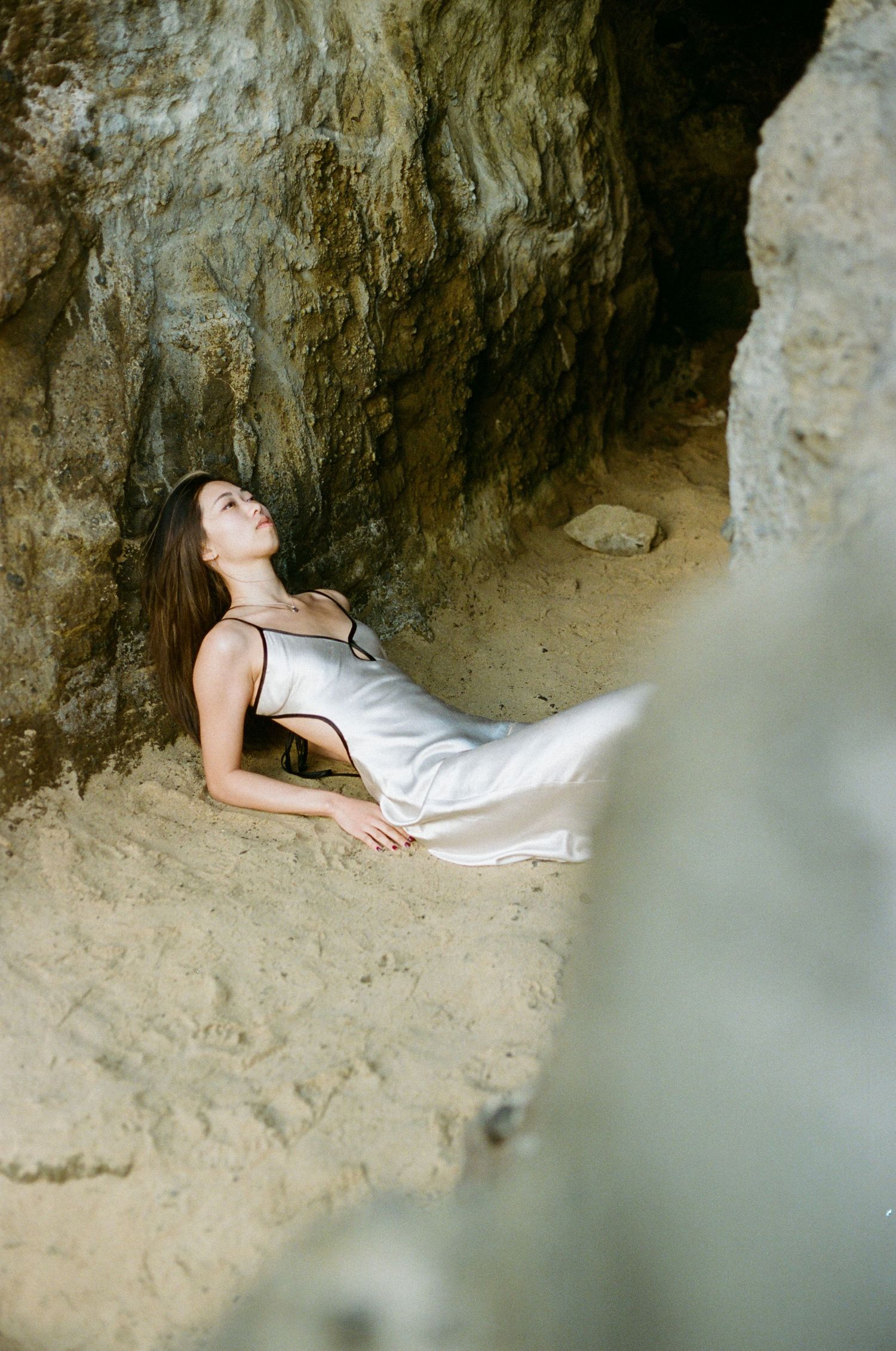 Woman in white slip dress reclining peacefully against rocky cave wall on sandy ground.