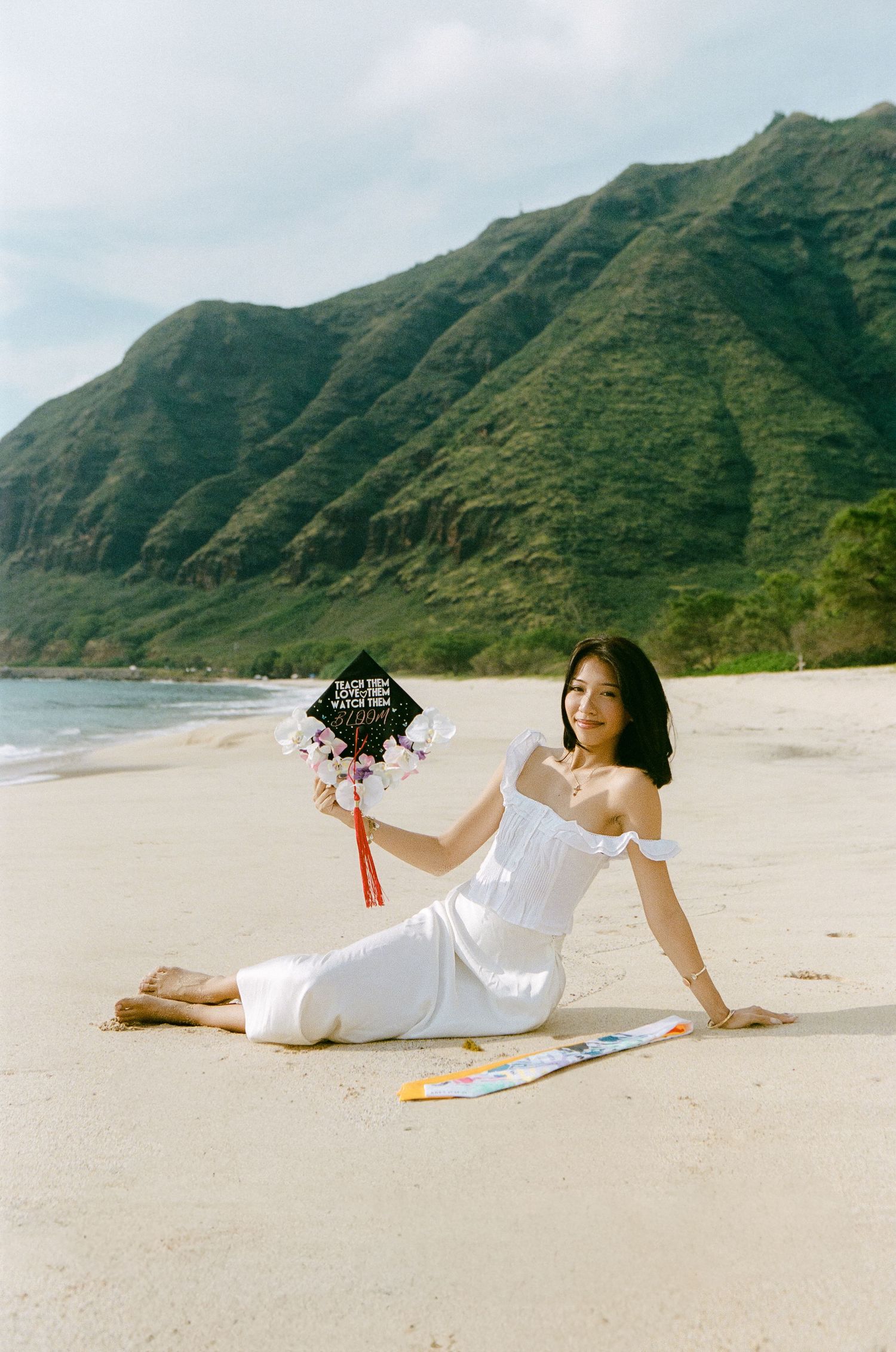 Graduate in white dress sitting on sandy beach holding decorated 2025 mortarboard with green mountains.