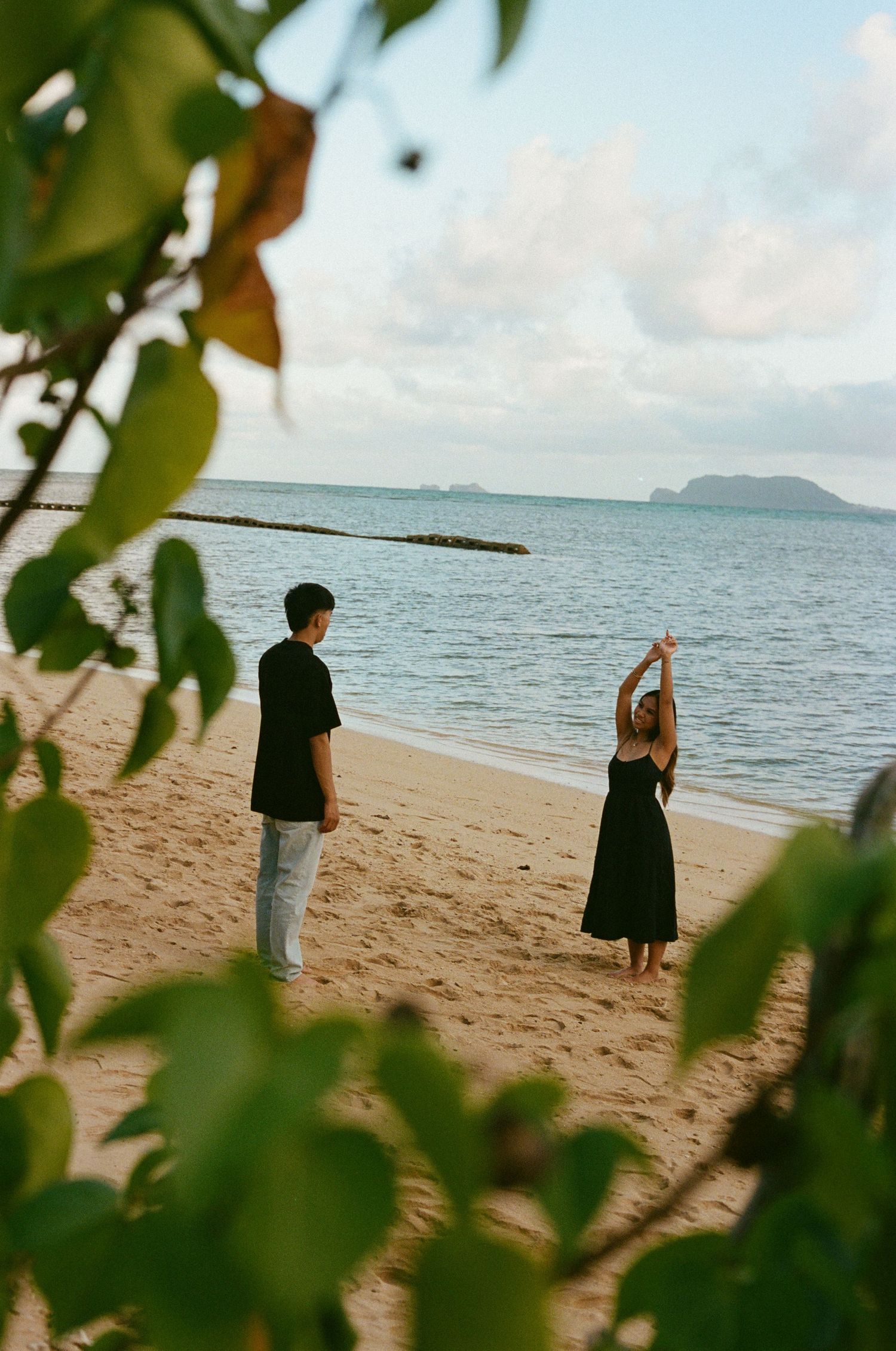Couple standing on sandy beach near ocean, woman with arms raised, framed by tropical foliage.