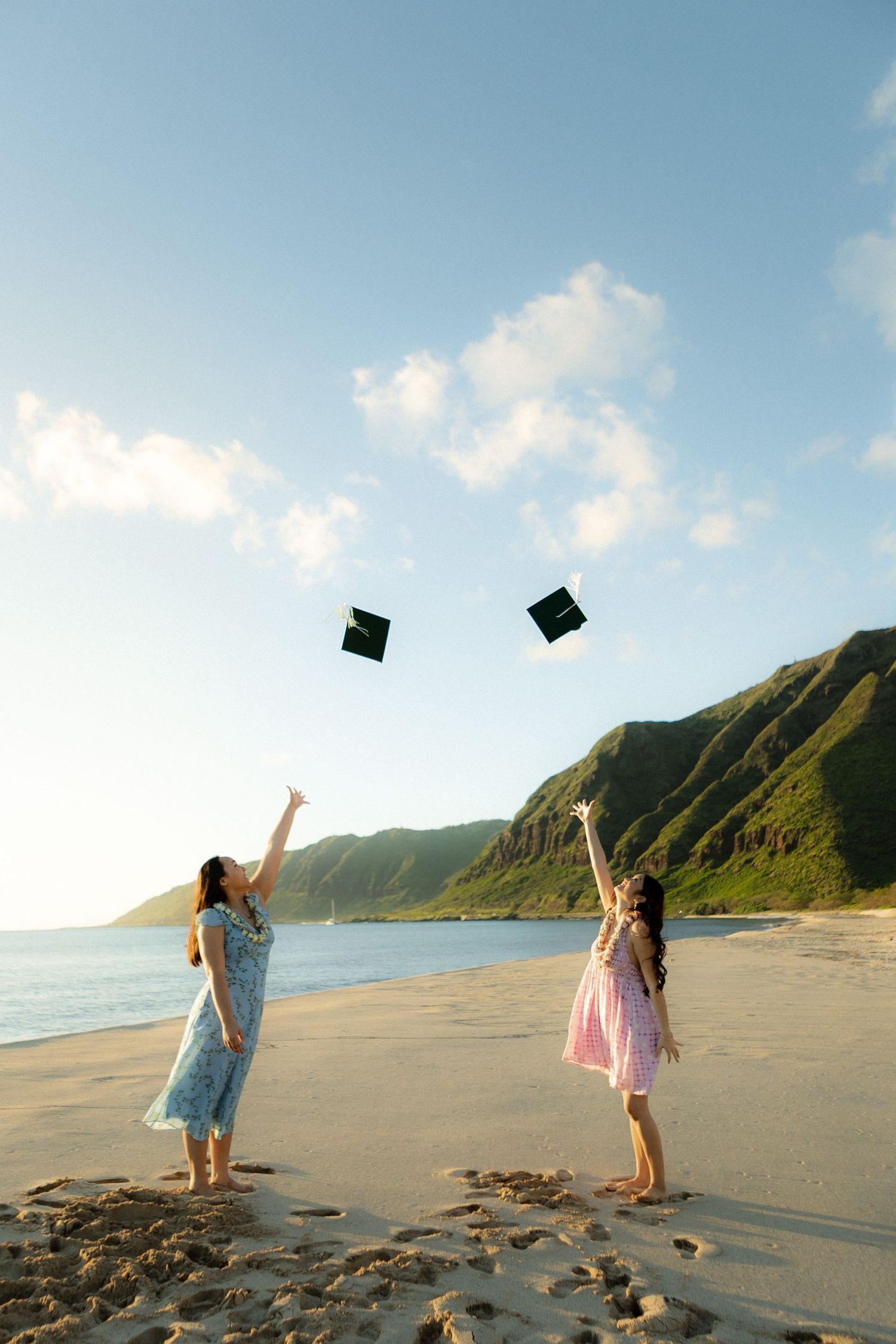 Two women toss graduation caps on a sandy Hawaiian beach with mountain backdrop.