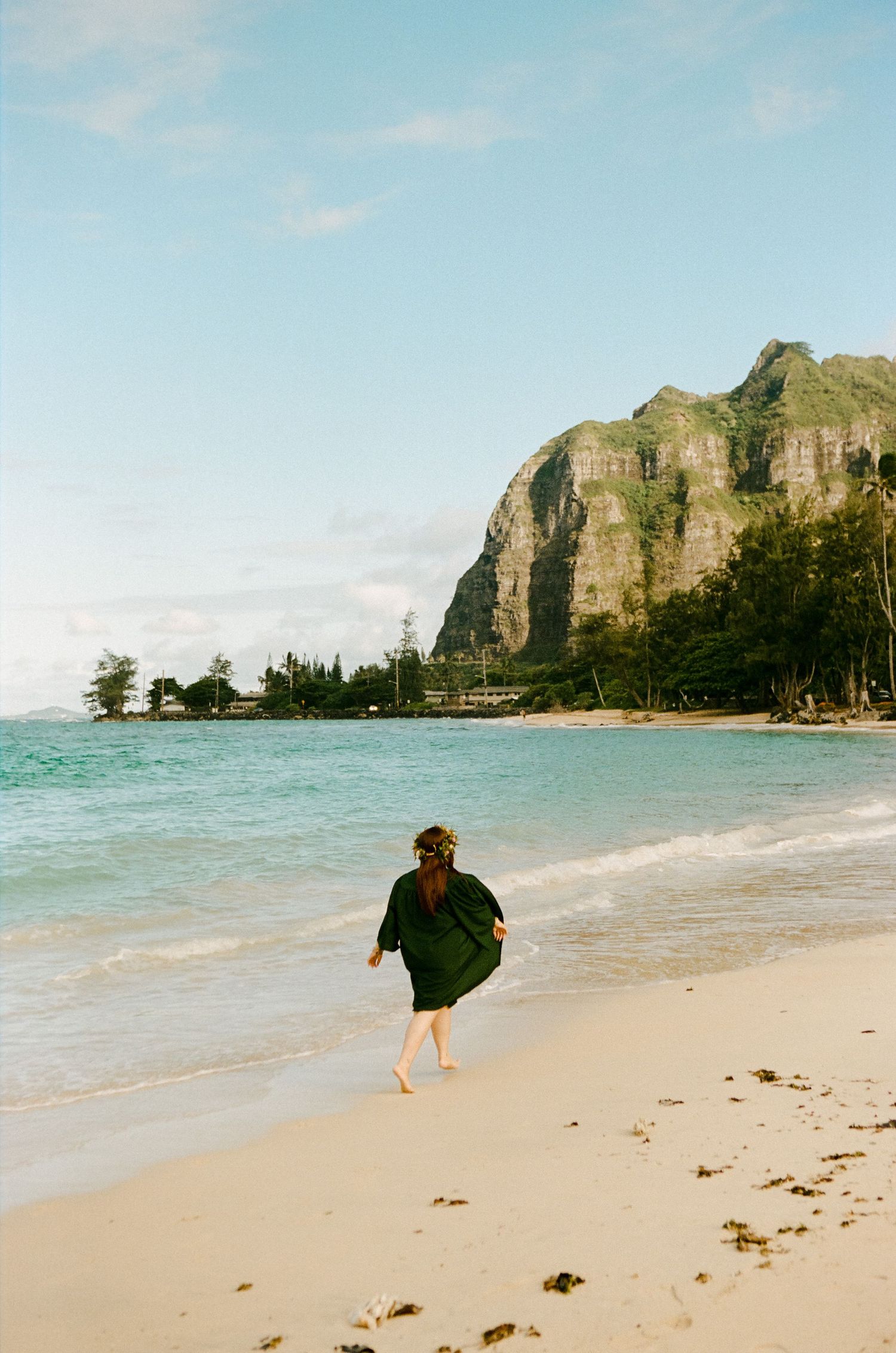 Woman in dark green dress walking barefoot along a scenic beach with dramatic cliffs in background.