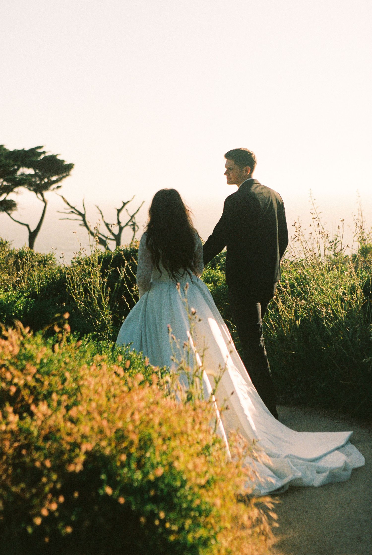 A couple in wedding attire holds hands on a coastal path at sunset, surrounded by lush greenery and a misty horizon.