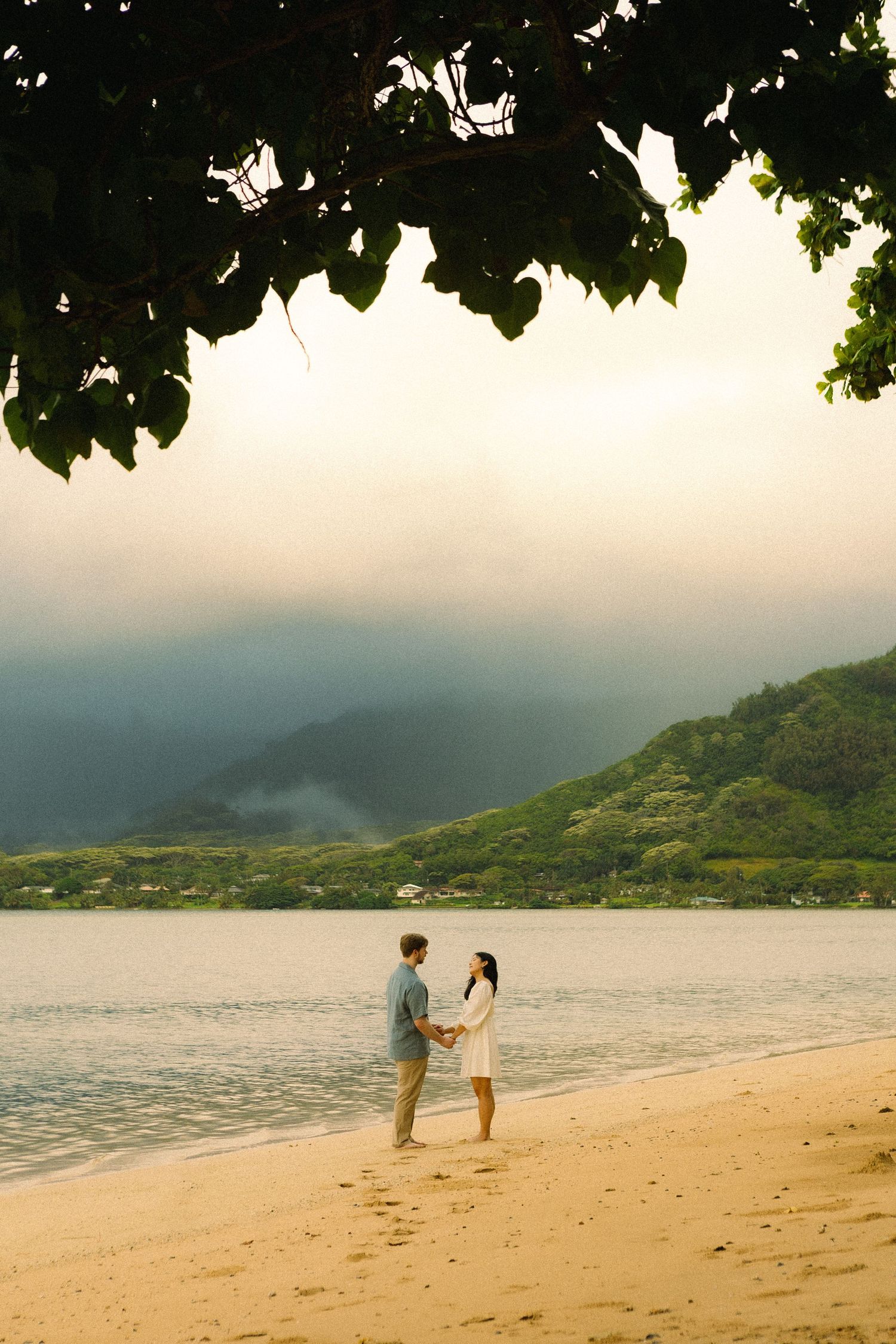 Couple holding hands on a tropical beach with lush green mountains and dramatic cloudy sky in background.