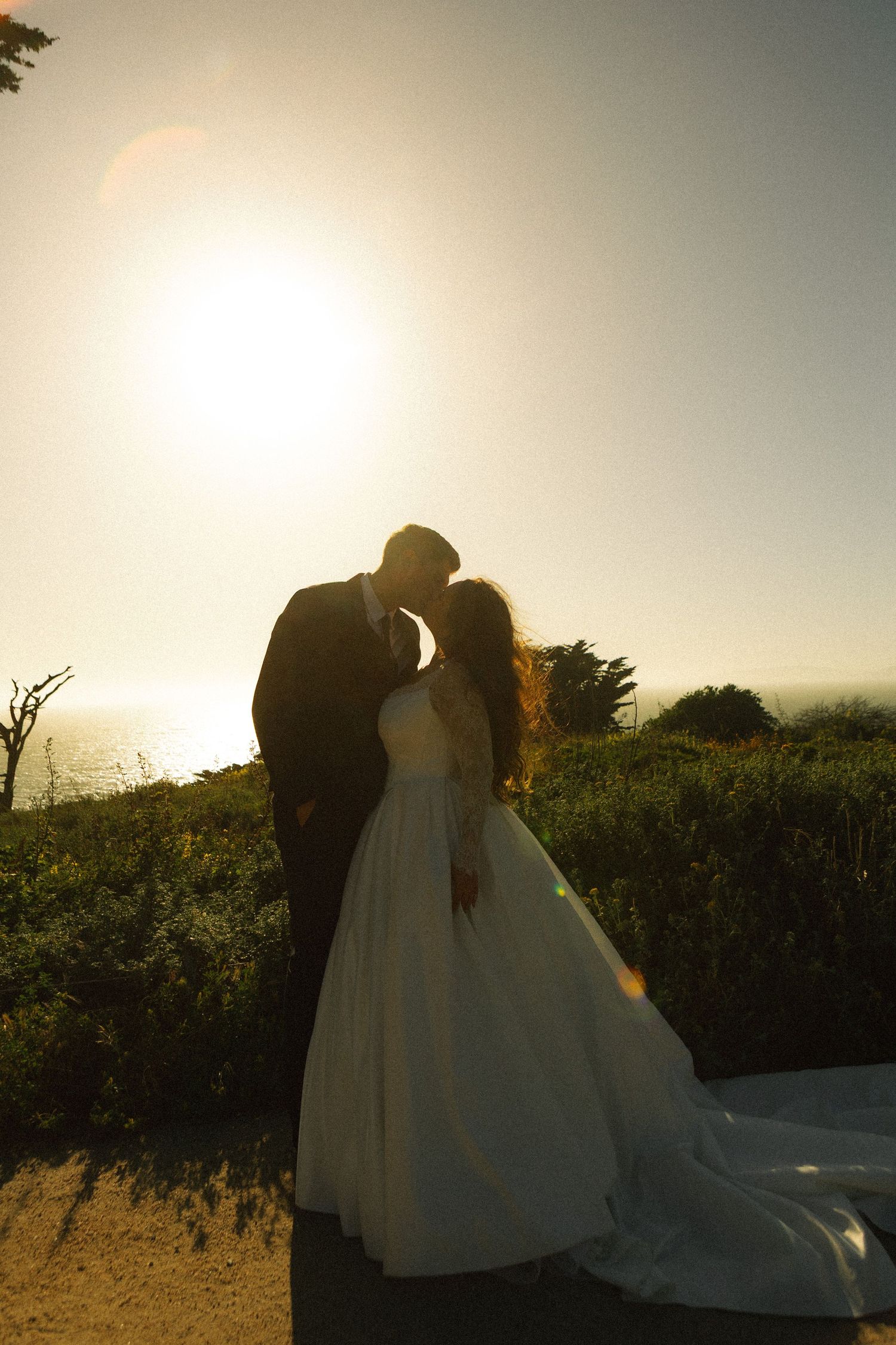 Bride and groom silhouetted against golden sunset with ocean views, sharing romantic moment in wedding attire.