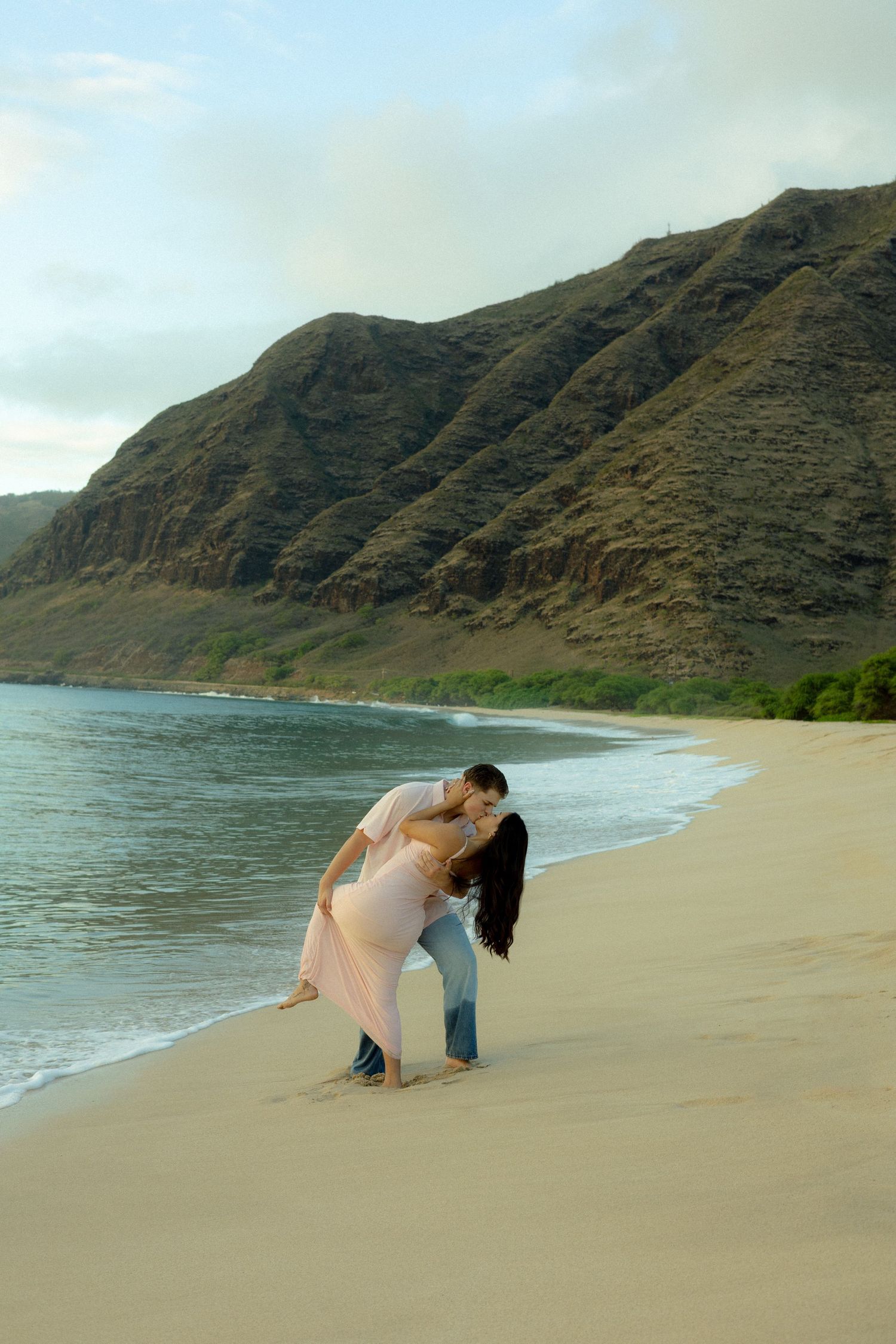 Couple sharing a romantic dip kiss on a serene Hawaiian beach with mountain backdrop.