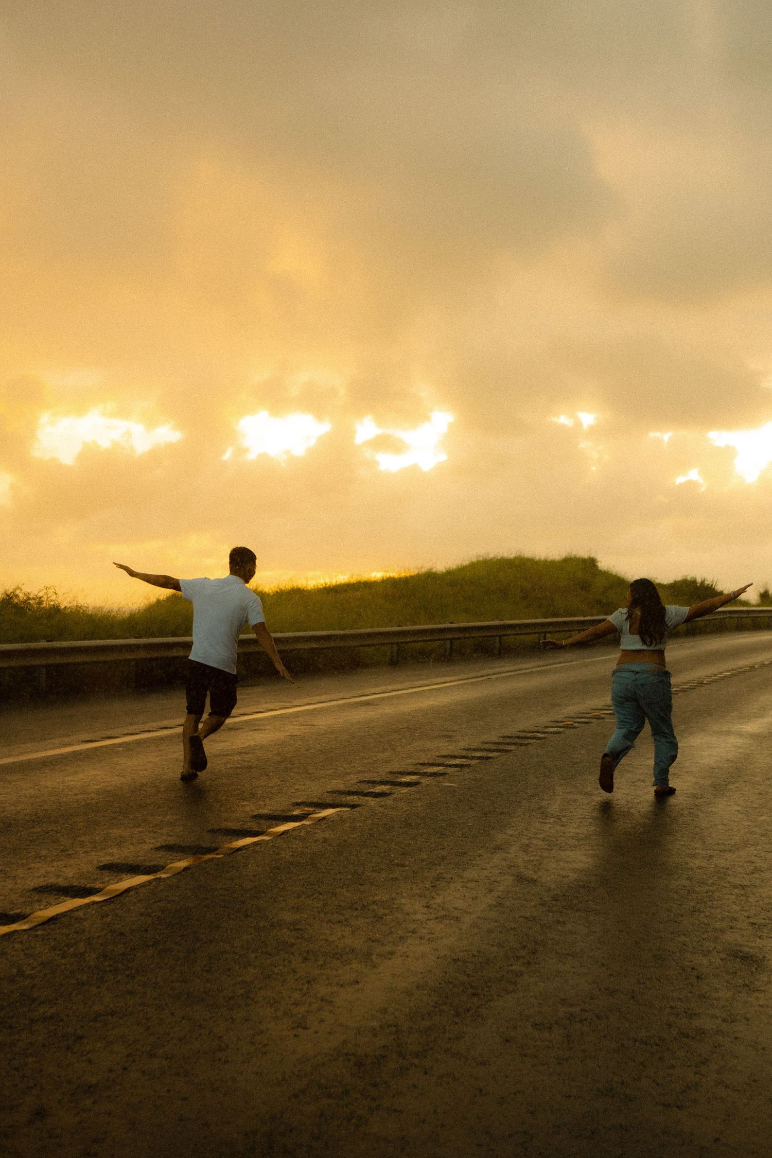 Two people running on a wet road at sunset with golden cloudy sky and green hills in the background.