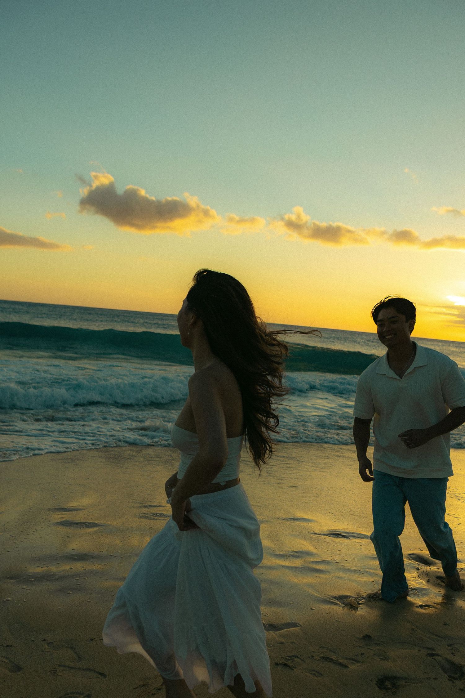 Couple on sandy beach at vibrant golden sunset, woman in white dress with flowing hair.