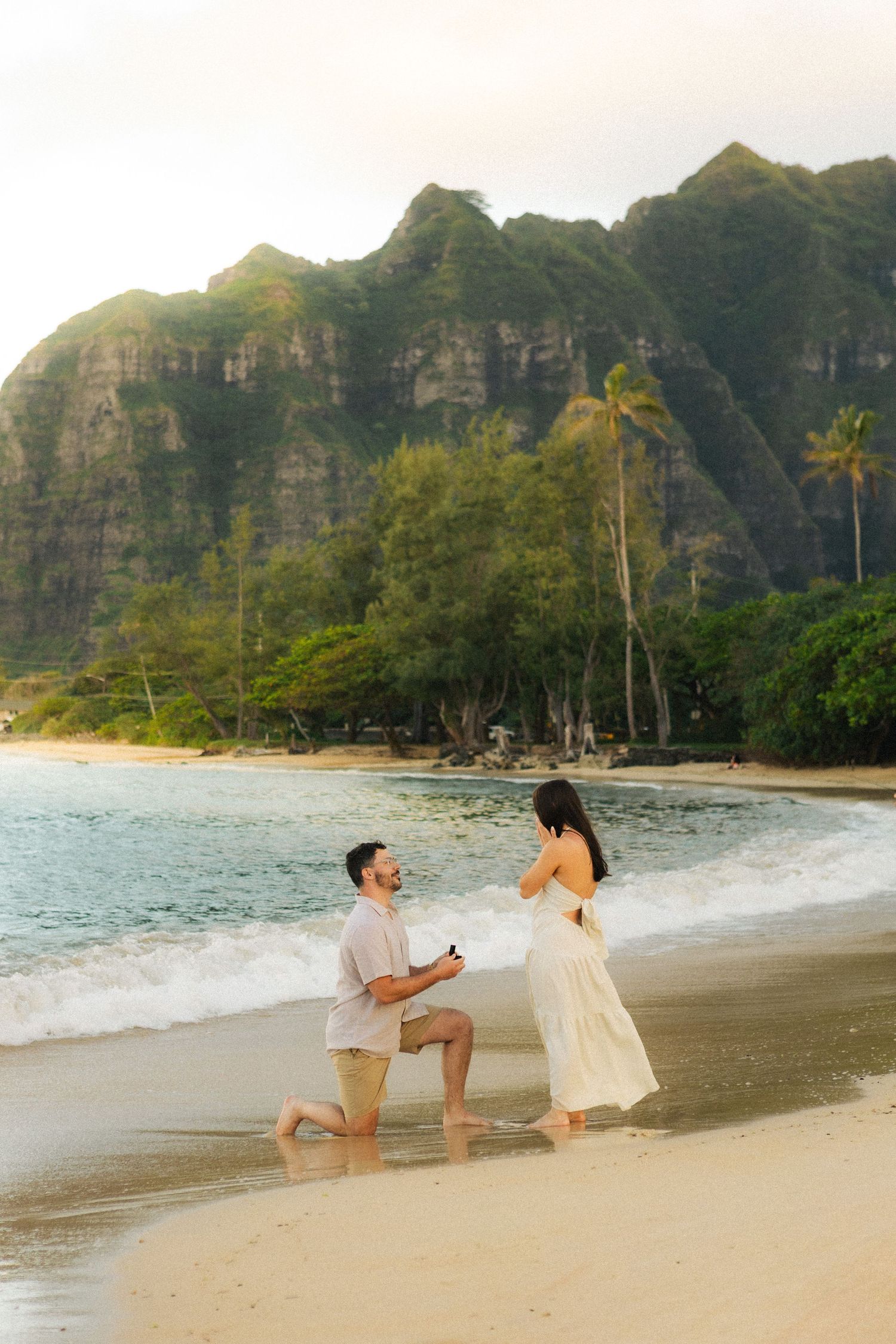 Man proposing on one knee to woman on Hawaiian beach with lush cliffs behind.