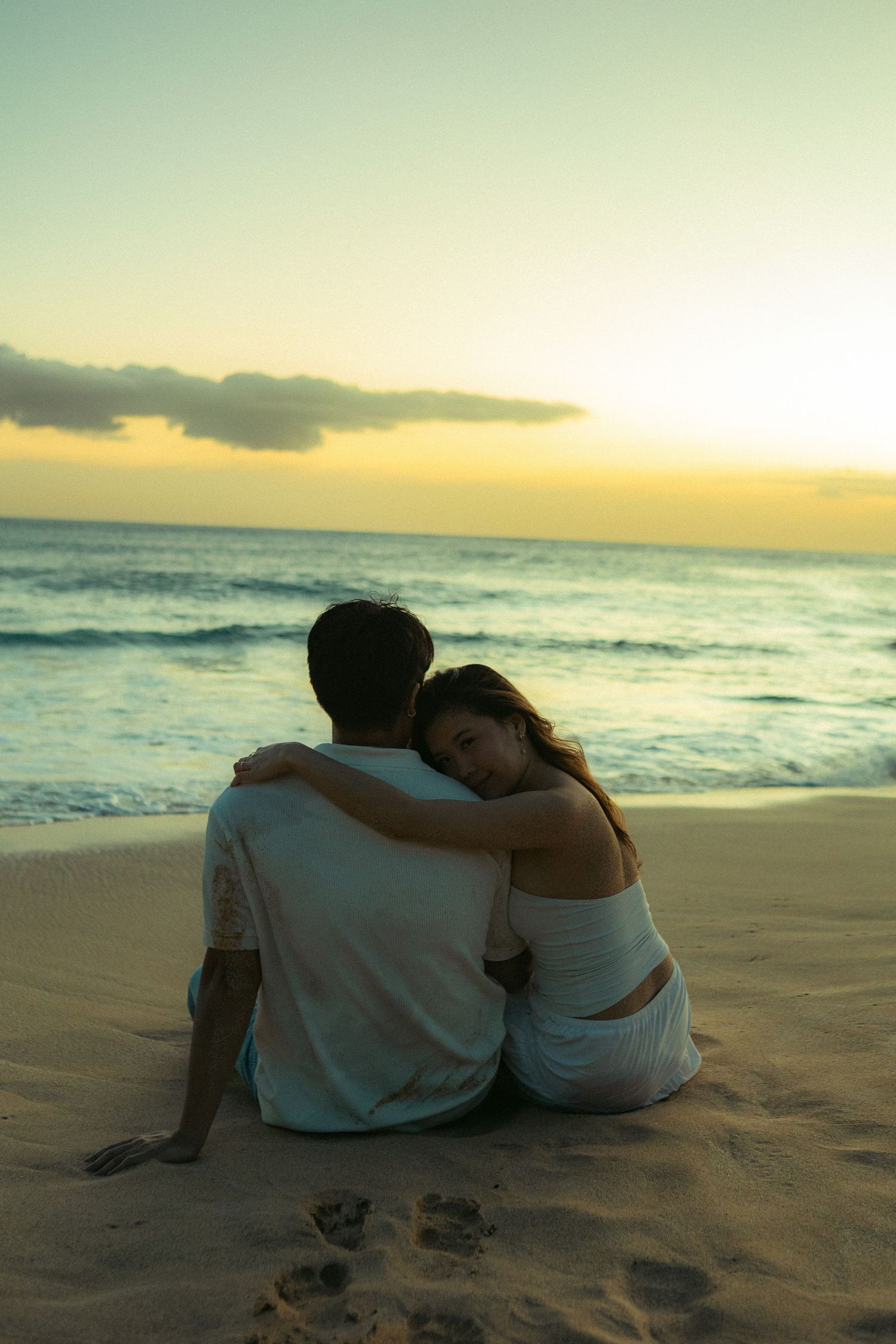 Romantic couple sitting together on beach sand at sunset, woman resting head on mans shoulder.