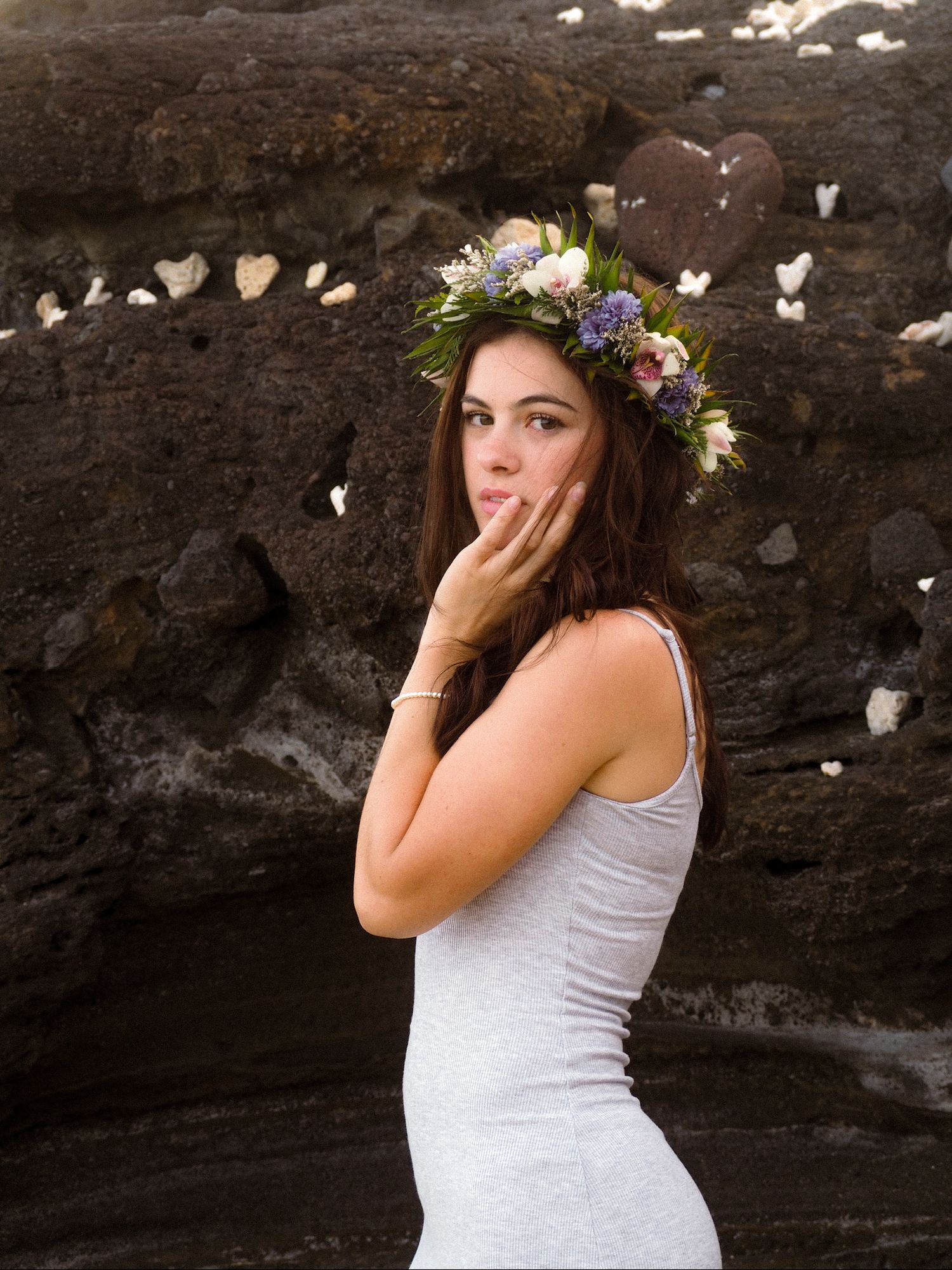 Woman in gray dress with colorful floral crown blowing a kiss near textured volcanic rock wall.