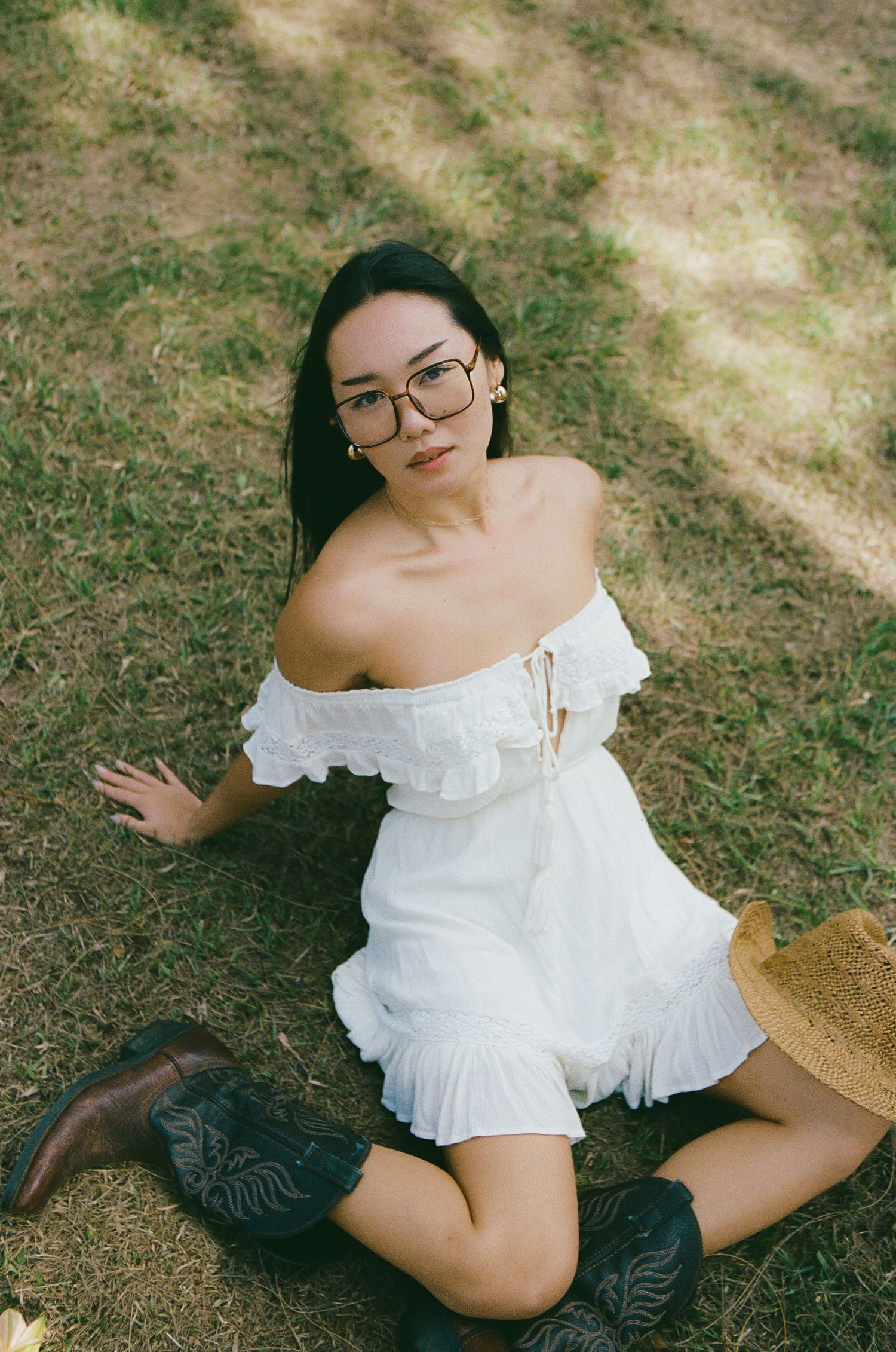 Young woman in white off-shoulder dress and cowboy boots sitting on grass with straw hat.