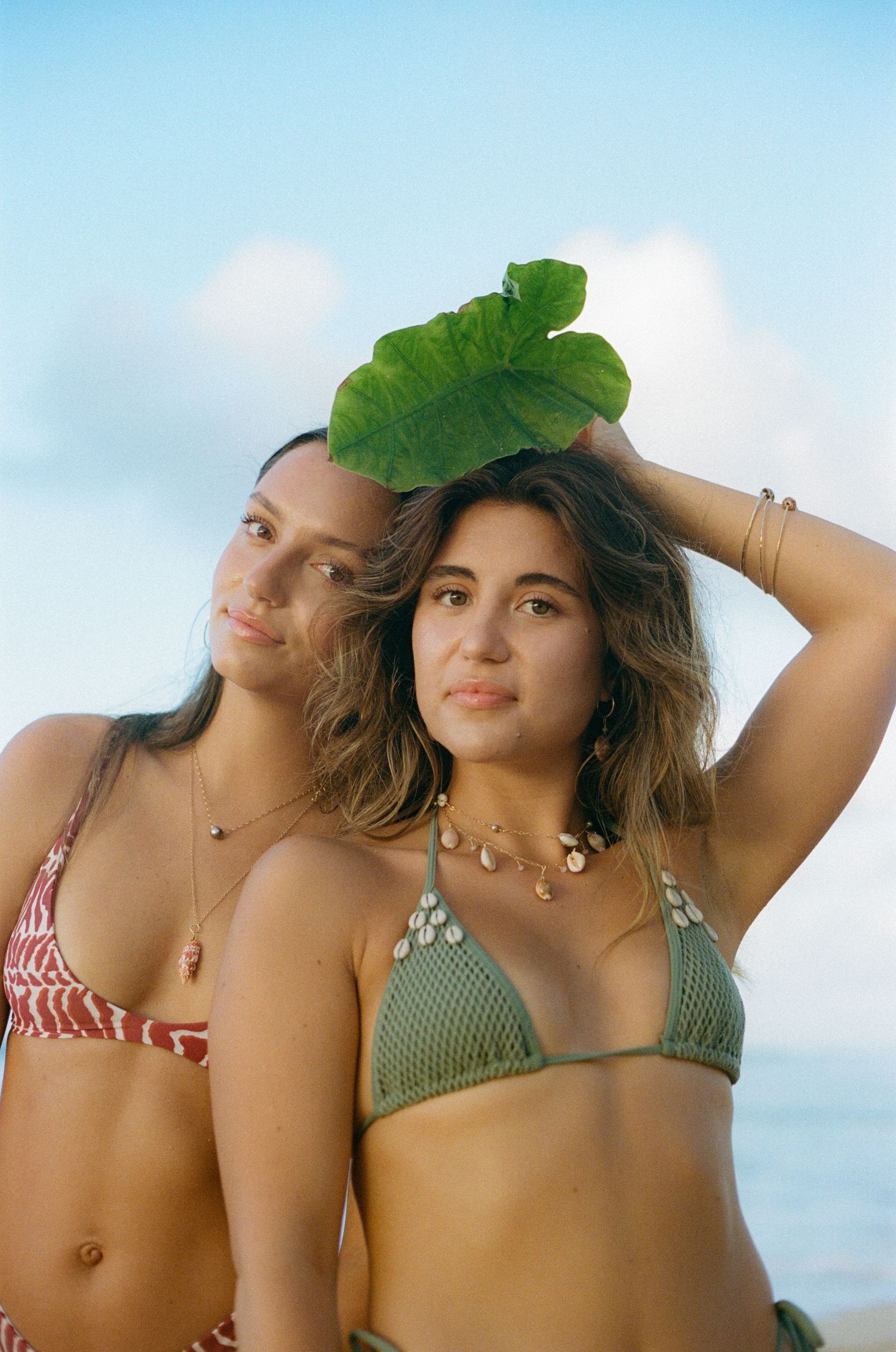 Two women in bikinis at the beach, one holding a large tropical leaf overhead.