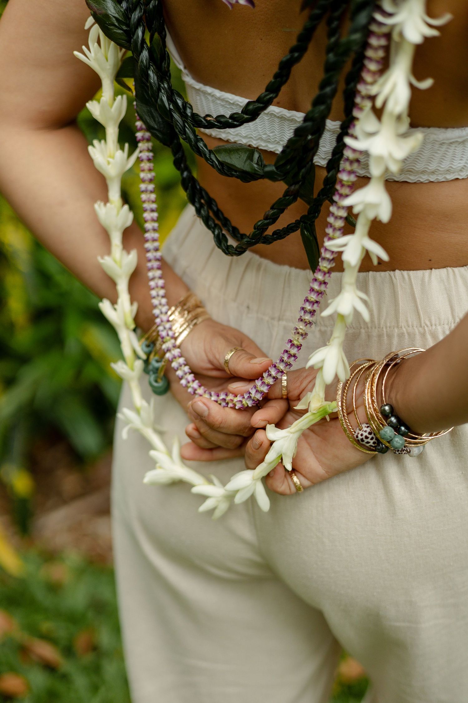 Close-up of hands holding white tuberose lei wearing gold bangles and beaded necklaces.