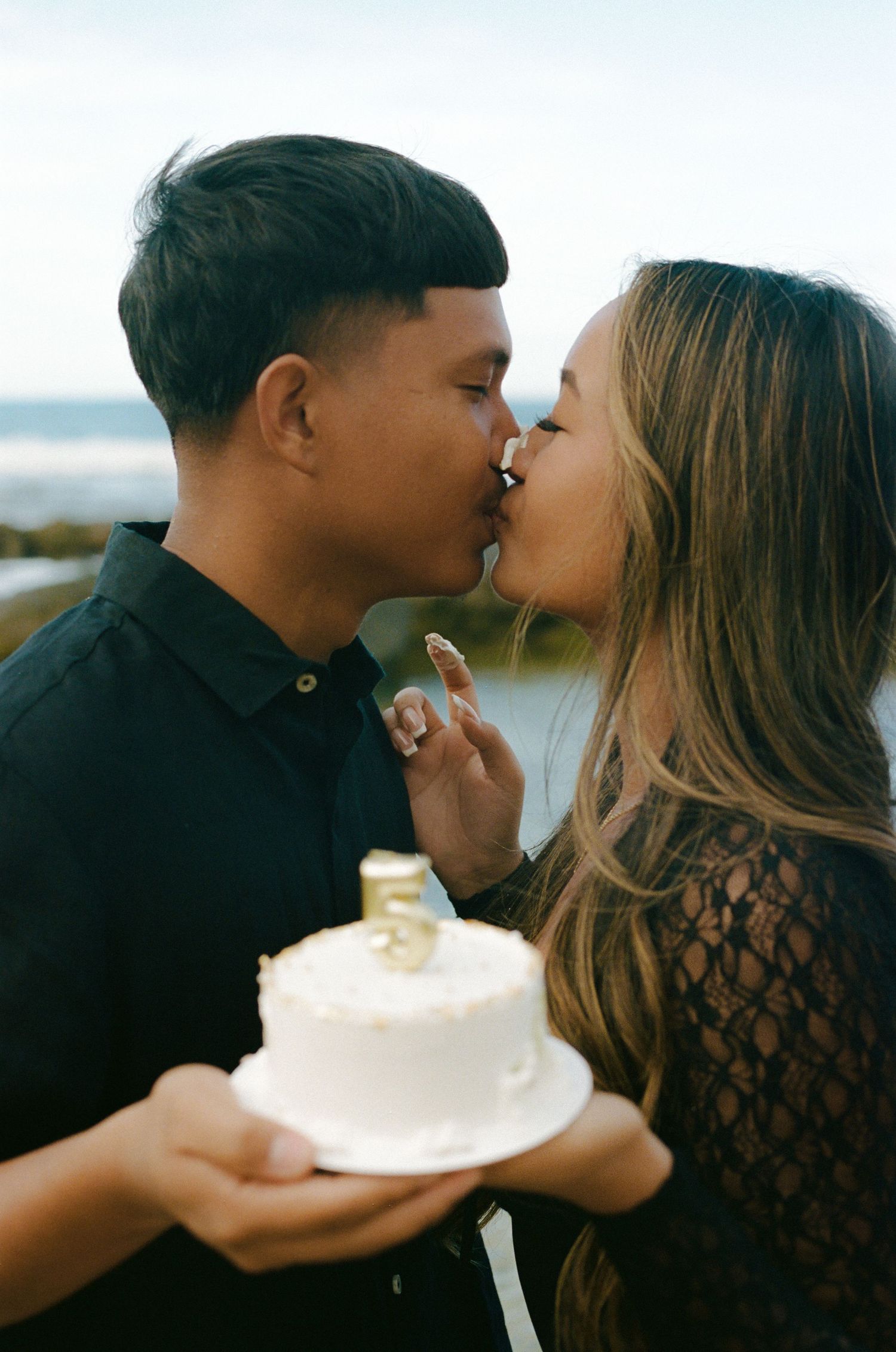 A couple shares a romantic kiss while holding a white anniversary cake near the ocean.